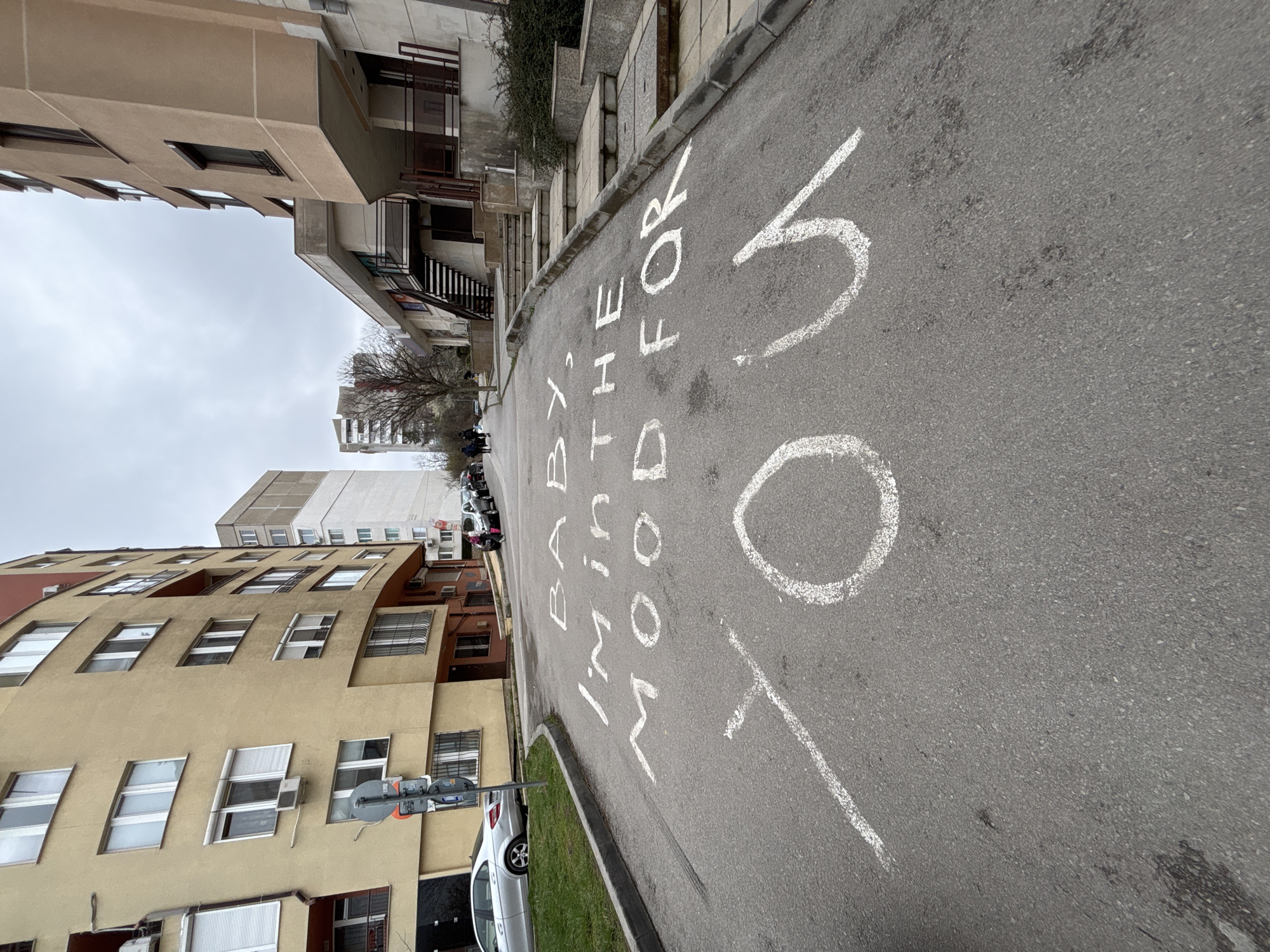 The image shows an urban street scene in Sofia, Bulgaria, on a cloudy day. The street is lined with buildings of varying architectural styles and heights, constructed from a mixture of light-colored concrete and brick. A two-way street has white painted messages that read "OUT FOR BODY AND SOUL" and "TO". Cars are parked along the side of the street, a traffic light is on the left. Bare trees are visible and several individuals are walking in the distance. The image has been rotated counterclockwise.