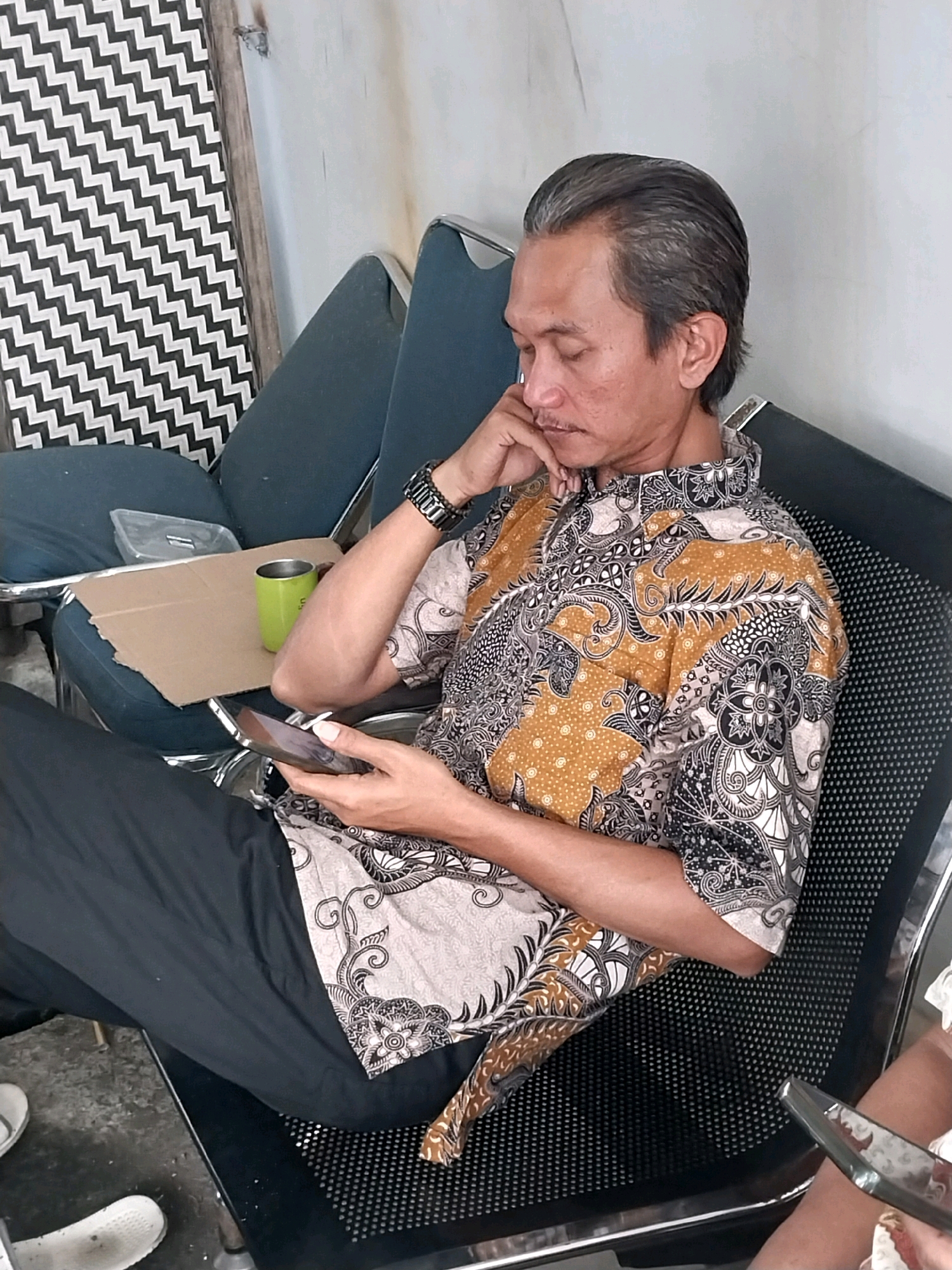 A man with graying hair sits in a chair, looking at his phone. He is wearing a batik shirt and black pants. He is holding his chin with his left hand. In front of him, on a cardboard piece, is a green mug and a clear plastic container. To his left is a wall with a black and white zigzag pattern. The man is in a room, possibly at an office or waiting area.
