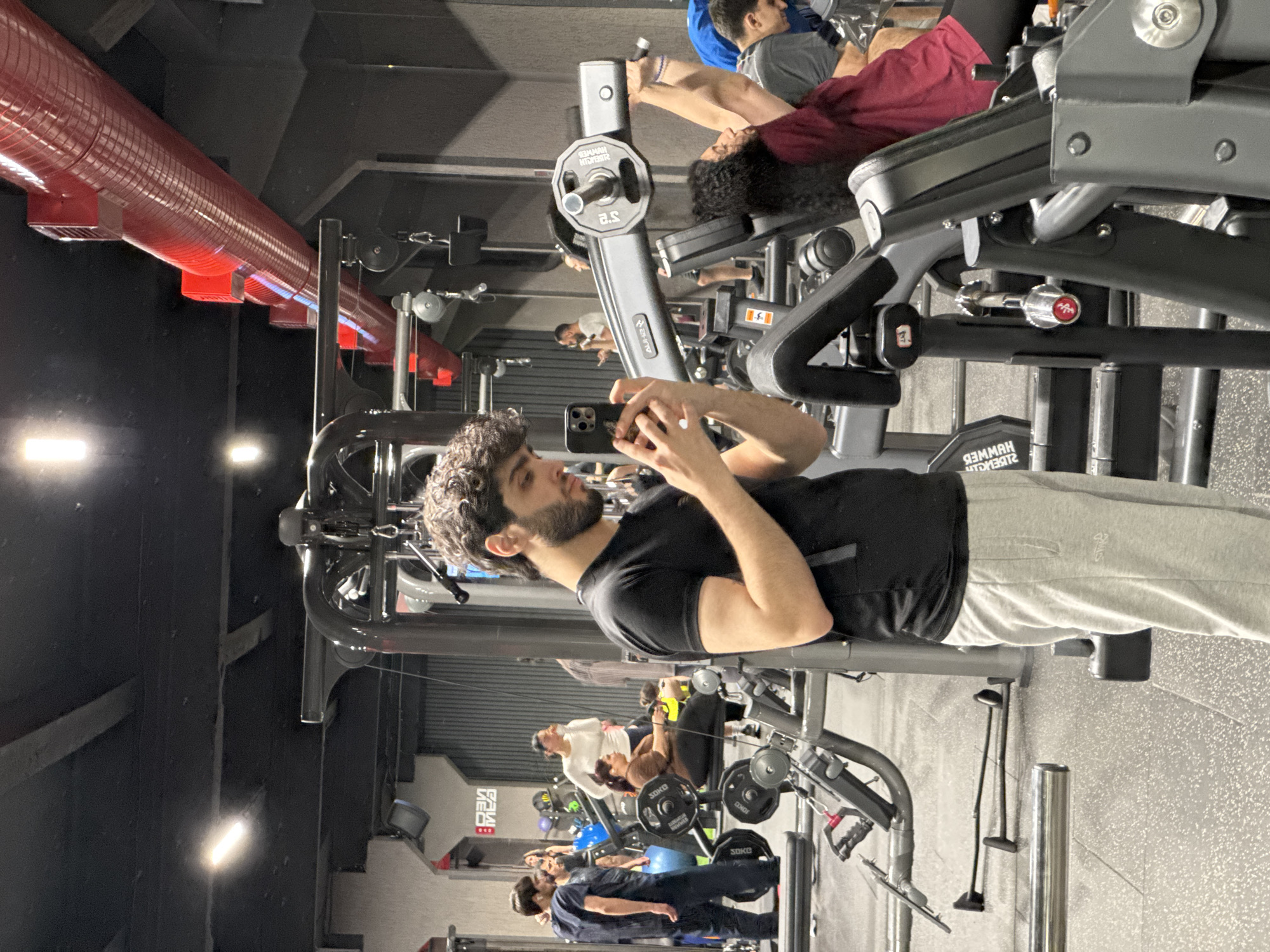 The image shows the interior of a gym, possibly located in Yerevan, Armenia, although this location is not discernible within the image itself. A man is lying on a weight machine, looking at his phone. He is wearing a black t-shirt and grey sweatpants. In the background, several other people are using gym equipment. There are various weight machines and gym apparatus visible. A red duct system is seen along the ceiling. The lighting is bright, and the floor appears to be tiled.
