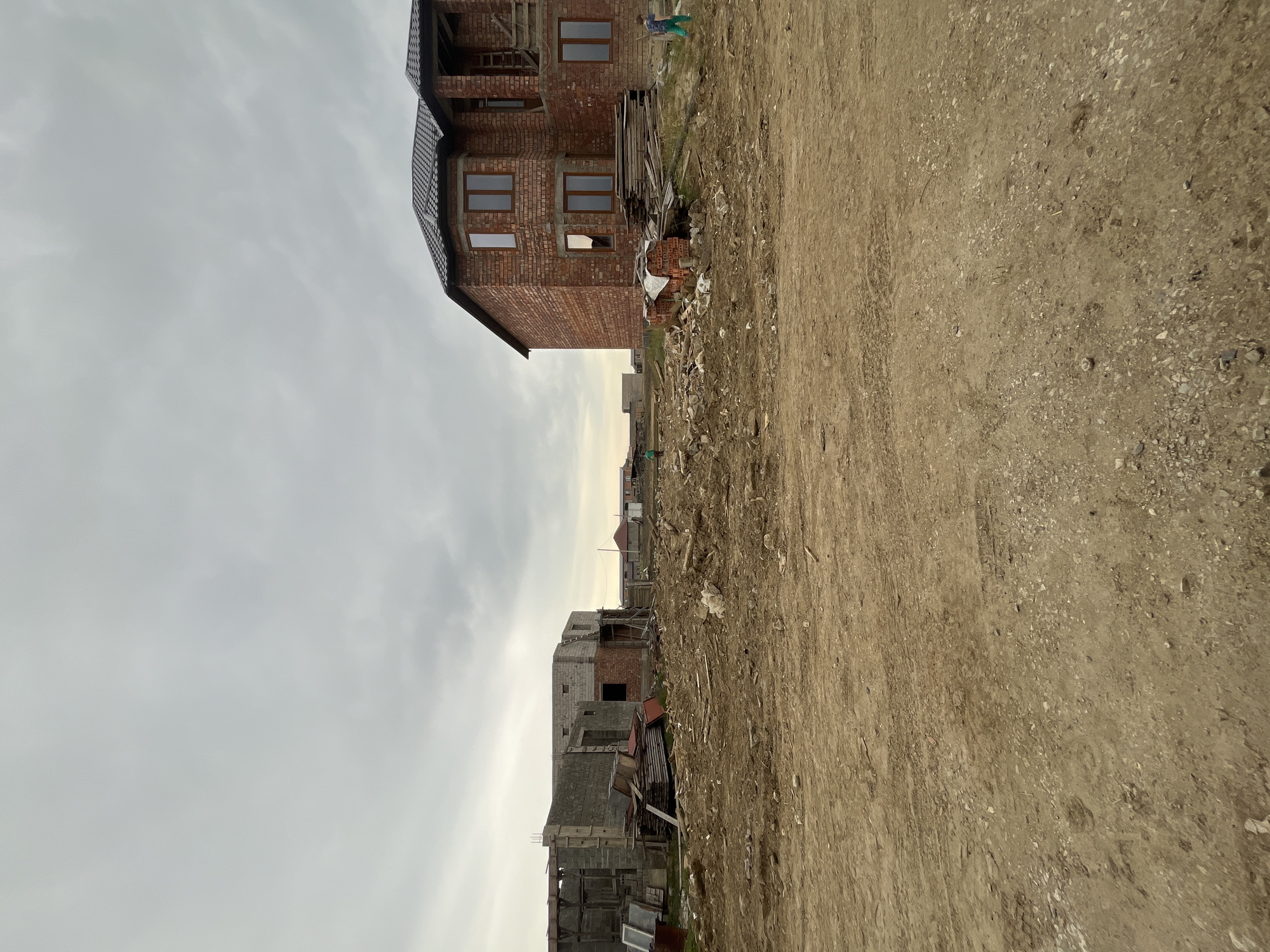 The image captures a residential construction zone in Kaspiysk, Russia, under an overcast sky. The foreground consists of a vast expanse of disturbed, light brown earth, marked by tire tracks and scattered debris.
On the left midground, an unfinished house structure is visible, built with grey concrete blocks and some red brickwork. This structure appears to be in an early stage of construction, with exposed framework and openings for future windows and doors. Piles of wooden planks, metal sheets, and discarded window frames are accumulated around it, consistent with construction waste and materials.
In the center midground, the bare earth continues, stretching towards the horizon where distant, low-rise structures are faintly visible. Two individuals are present: one person wearing green clothing walks towards the left in the distance, while another, in green pants and a blue top, walks towards the right, closer to the more complete house. The latter individual appears to be carrying a long object.
On the right midground, a more developed two-story house stands, constructed from reddish-brown brick with a dark, pitched roof. It features multiple installed windows with dark frames. An unfinished section on the right side of this house shows additional brickwork and a wooden ladder leaning against it, indicating ongoing work. A stack of new red bricks, partially covered by a white tarp, and additional wooden planks are positioned near the base of this structure.
The overall environment is characterized by undeveloped land and active building sites, under a uniformly grey, cloudy sky.