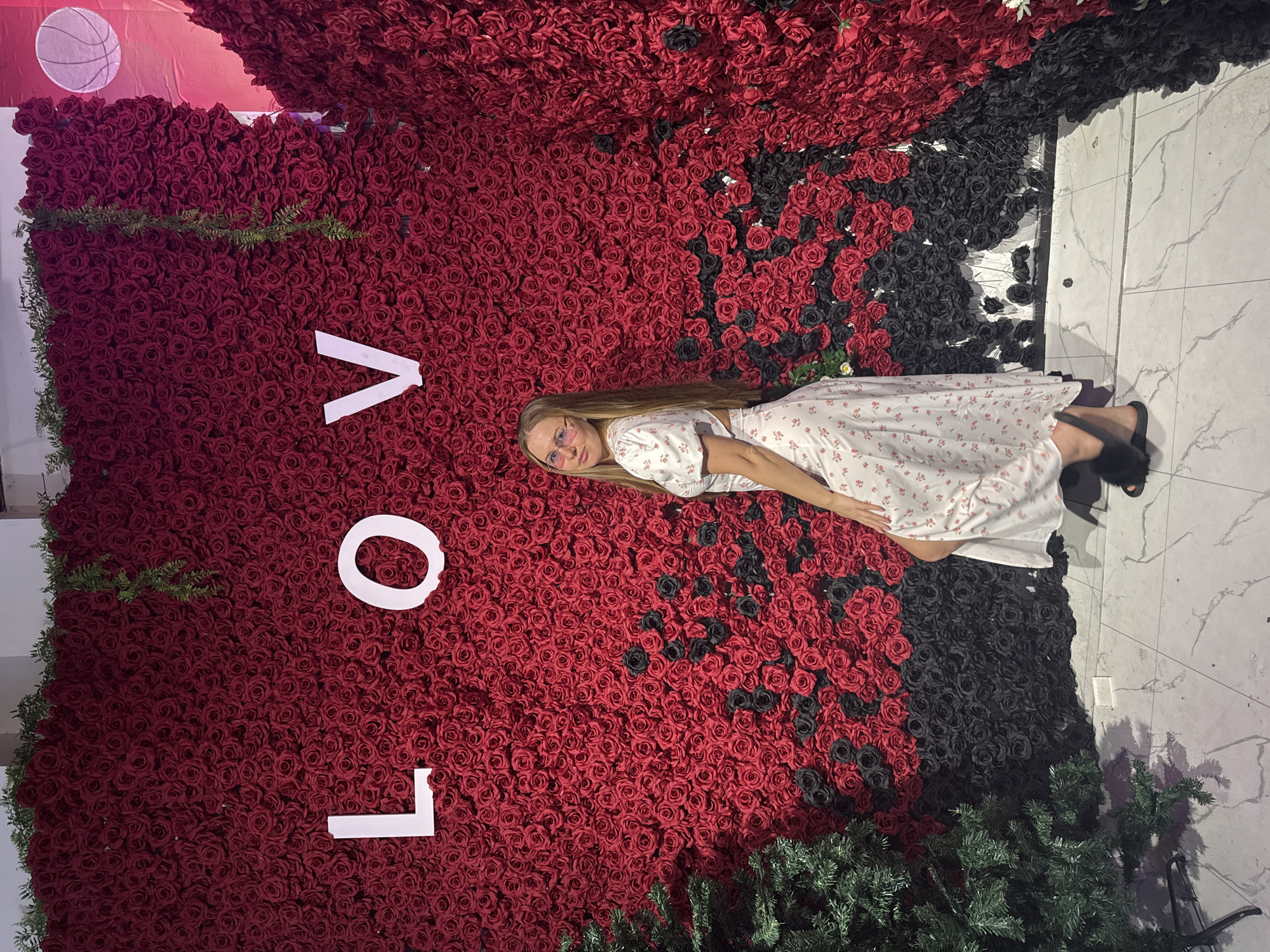 A young Caucasian woman with long blonde hair, wearing glasses and a white floral dress, stands in front of a large wall of red roses. The wall has white letters spelling "LOVE" on it, and a green artificial Christmas tree stands to the left. The floor is tiled with a marble pattern. The backdrop appears to be a commercial space, possibly a photo studio or event venue, in Yiwu, China, a city known for its wholesale markets. The overall lighting suggests an indoor environment, likely with artificial illumination.