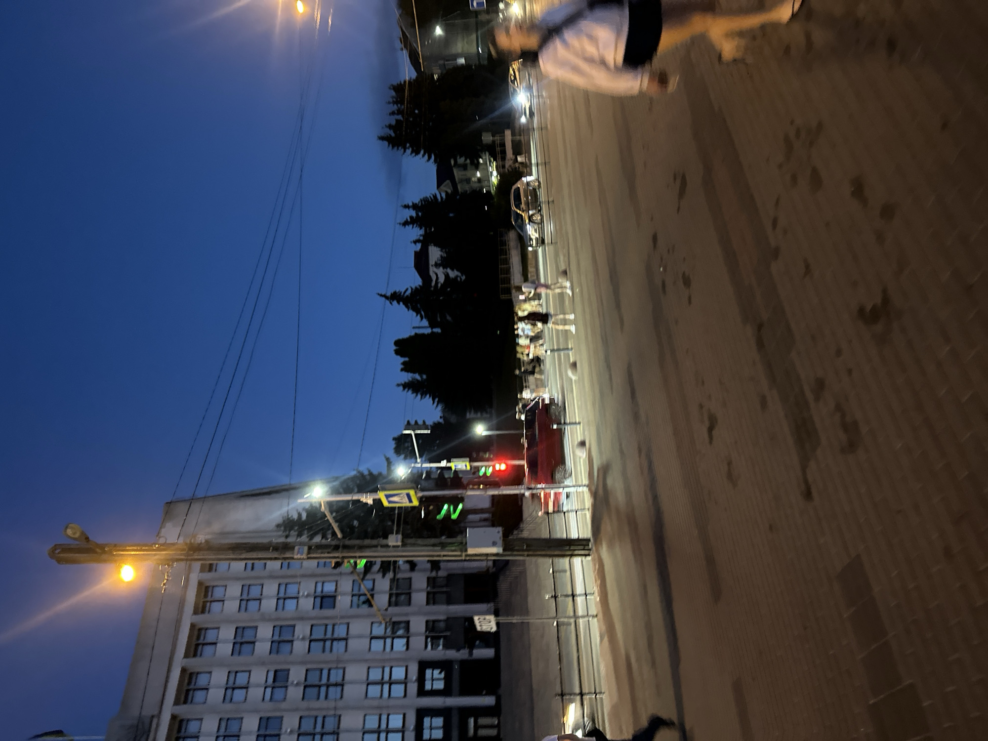 The image depicts a street scene at dusk or early evening in Ternopil, Ukraine, with a prominent building on the left and a street with pedestrian and vehicular traffic. The sky is a deep blue, indicating twilight. A large, multi-story building with numerous windows, likely an administrative or institutional structure, dominates the left side of the frame. Its facade appears to be light-colored concrete or stone.
A street light with a bright yellow bulb illuminates the area, casting a warm glow. Traffic lights are visible, with a red light illuminated on one of them, suggesting that vehicles are stopped. A crosswalk is marked on the street. Several people are crossing the street or walking on the sidewalk. One individual in the foreground, on the right side of the image, is walking towards the viewer and is slightly out of focus. This person is wearing a light-colored top and dark shorts or skirt. Other individuals, appearing as silhouettes or less defined figures, are visible further down the street, some near the crosswalk.
Vehicles are present on the road. A red car is stopped near the crosswalk, and other cars are visible further down the street, some with their headlights on. The street surface appears to be paved with brick or cobblestone, with some wet patches or reflections visible, indicating recent rain or a damp surface. Trees are silhouetted against the sky in the background, and power lines are strung across the scene. A "STOP" sign is mounted on a pole near the building on the left. The overall impression is of a busy urban intersection during a transitional period of the day.