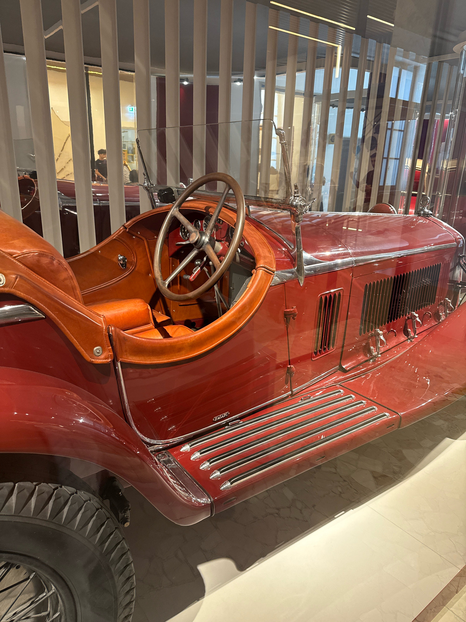 The image shows a close-up, eye-level view of a vintage red car, possibly in a museum setting in Milan, Italy. The car features a polished red exterior and rich tan leather interior, including the seats and steering wheel. Details like the spokes of the steering wheel, chrome accents, and louvers on the side of the car are visible. The car is positioned behind a reflective barrier, possibly glass, which distorts the background slightly. In the distorted background, through a vertical slatted screen, the blurry figures of at least two people can be seen. The floor appears to be tiled with a marble pattern.