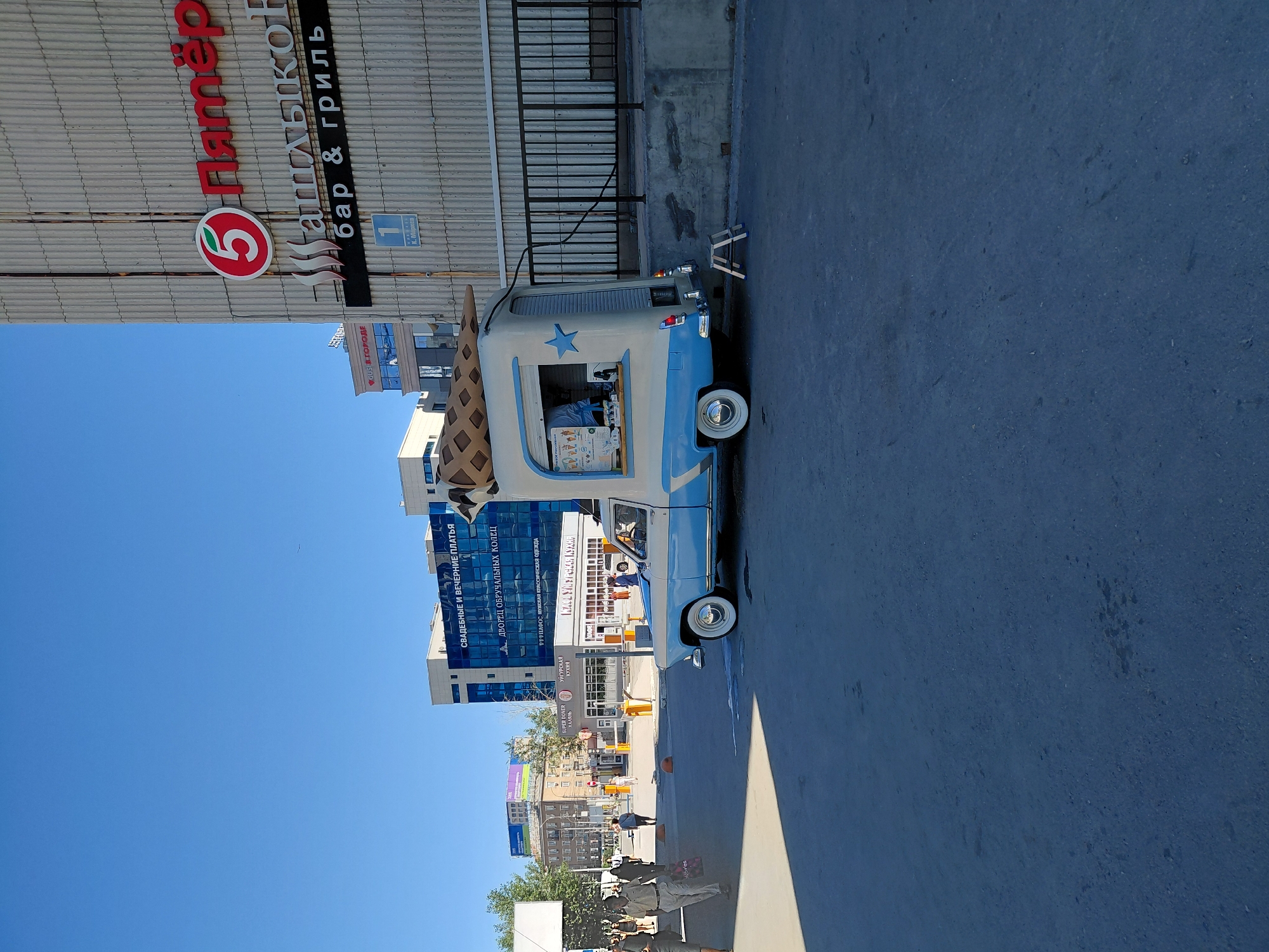 An outdoor scene on a sunny day shows a light blue and white retro-style ice cream truck parked on asphalt. The truck has a large waffle cone sculpture on its roof and a window where an employee, wearing a blue shirt, serves customers. In the background, there is a multi-story building with a "5 Пятёрочка" (Pyaterochka) supermarket sign, and another building with signs for "СВАДЕБНЫЕ И ВЕЧЕРНИЕ ПЛАТЬЯ" (Wedding and Evening Dresses) and "ДВОРЕЦ ОБРУЧАЛЬНЫХ КОЛЕЦ" (Palace of Wedding Rings). Several people are walking in the vicinity, and there is a black metal fence in the mid-ground. The overall environment appears to be a public urban space.