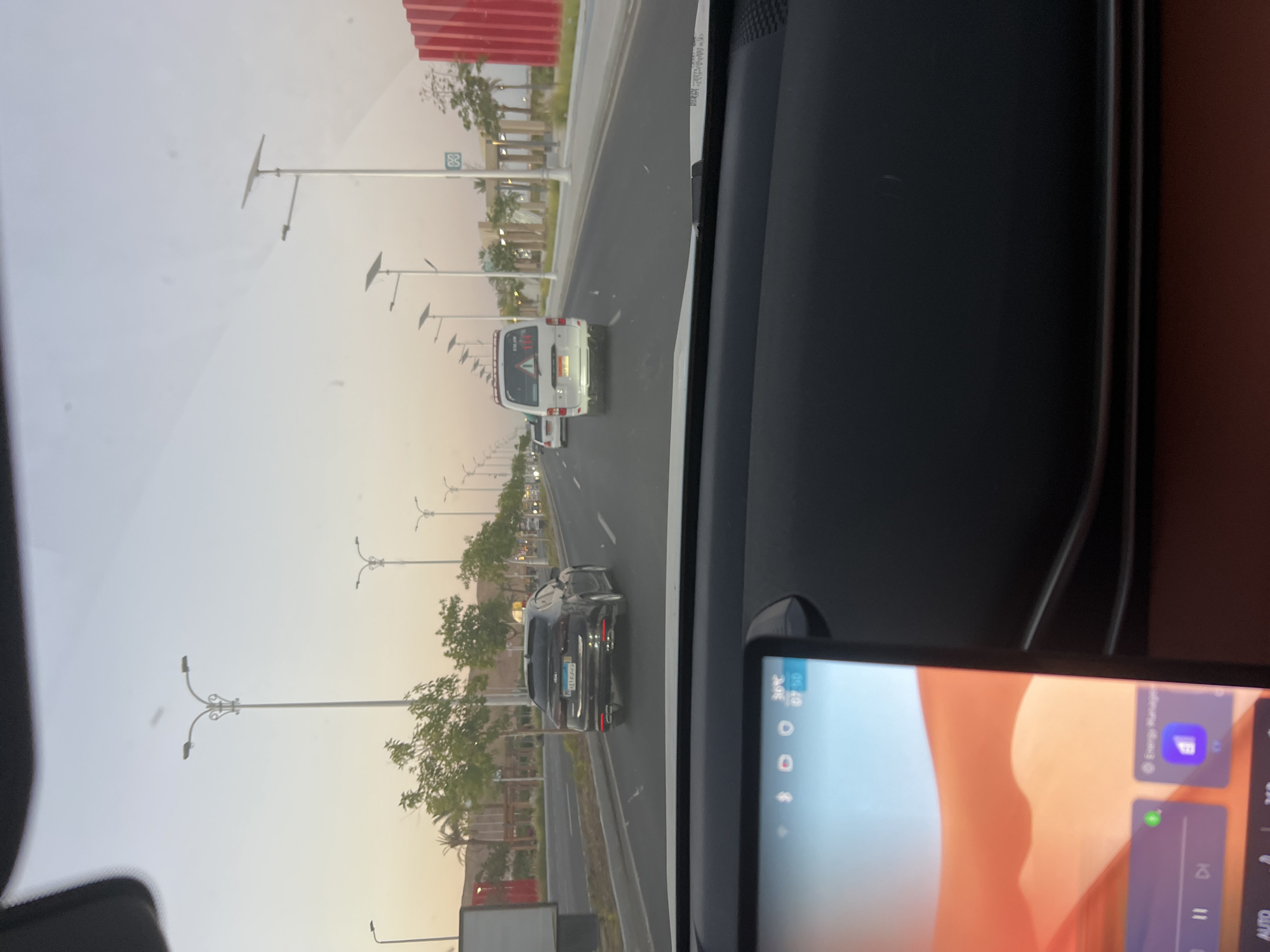 The image depicts a view from the driver's seat of a car in Sharm El Sheikh, Egypt, looking forward along a wide, multi-lane road. The time of day appears to be late afternoon or early evening, with a soft, dusky sky.
In the foreground, the car's dashboard and a large, modern touchscreen display are visible. The display shows what appears to be a custom interface with icons related to energy management and playback controls. The ambient temperature is displayed as 36°C.
On the road ahead, traffic is moving. The first vehicle in the middle lane is a black sedan with a yellow taxi sign on its roof. Behind it, in the right lane, is a white van, possibly a commercial vehicle or shuttle, with hazard lights illuminated and a red triangle warning sign on its rear window. Further down the road, other vehicles and streetlights are visible.
The roadside features modern urban infrastructure. Tall, slender lampposts, some equipped with solar panels, line the road. There are also paved sidewalks, decorative wooden structures, and greenery, including palm trees and other shrubs. In the distance, the architecture suggests a developed area, possibly a commercial or residential district. A striking, bright red, corrugated structure is visible on the right side of the road in the background, adding a strong visual element to the scene. The overall impression is of a well-maintained and contemporary urban environment in Sharm El Sheikh.