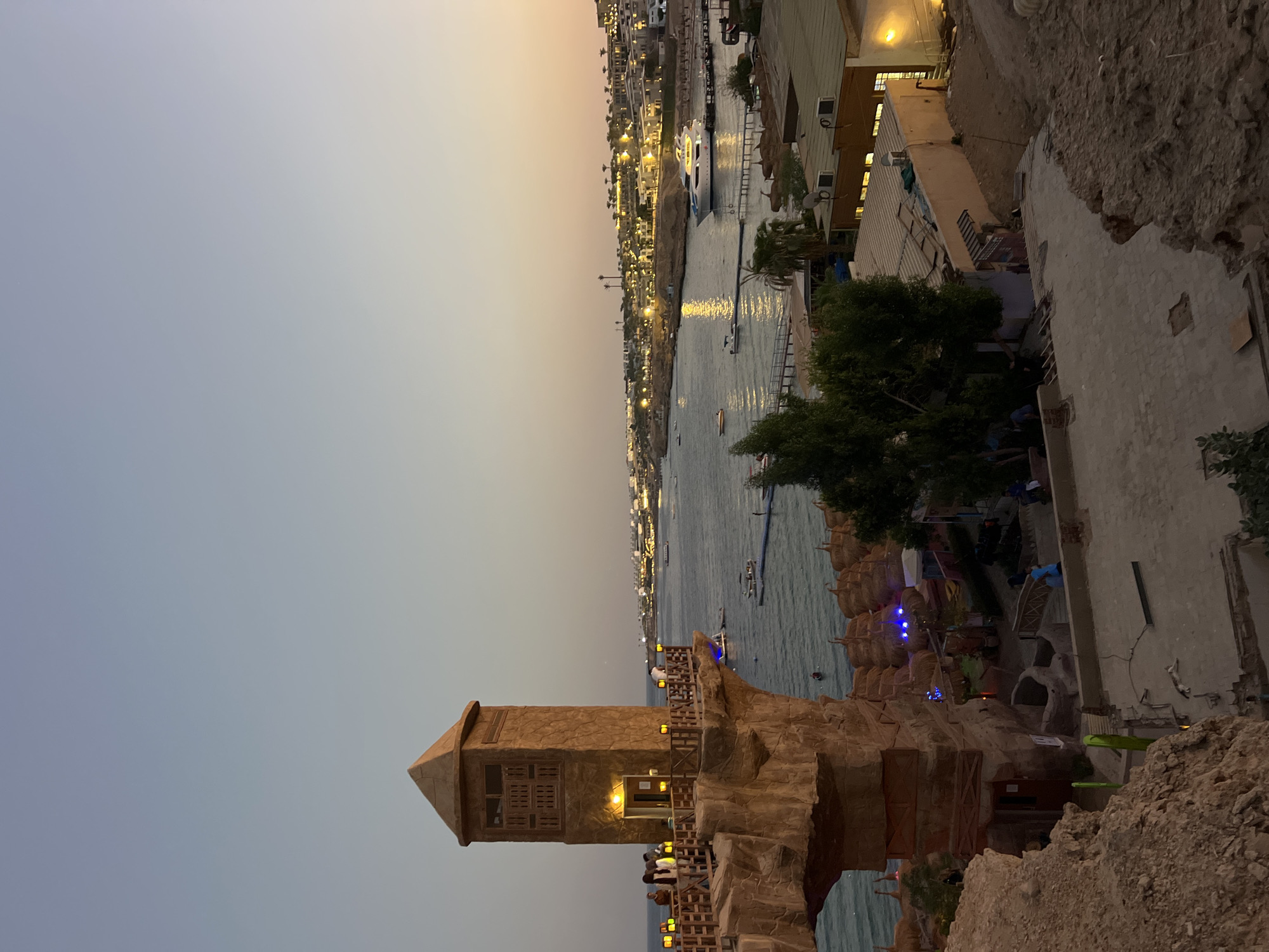 The image depicts a scenic view of the Red Sea coast in Sharm El Sheikh, Egypt, likely taken at dusk. In the foreground, a distinctive rock formation with a tower-like structure and a wooden balcony juts out over the water. Several people are visible on the balcony, some appearing to be tourists. The sea below is calm, with several boats and small vessels scattered across its surface. A town or resort area lines the coastline in the distance, with buildings illuminated by lights, suggesting evening activity. The sky transitions from a pale blue at the top to a warm, golden hue near the horizon, indicating either sunrise or sunset. The overall scene conveys a sense of relaxed vacation atmosphere.