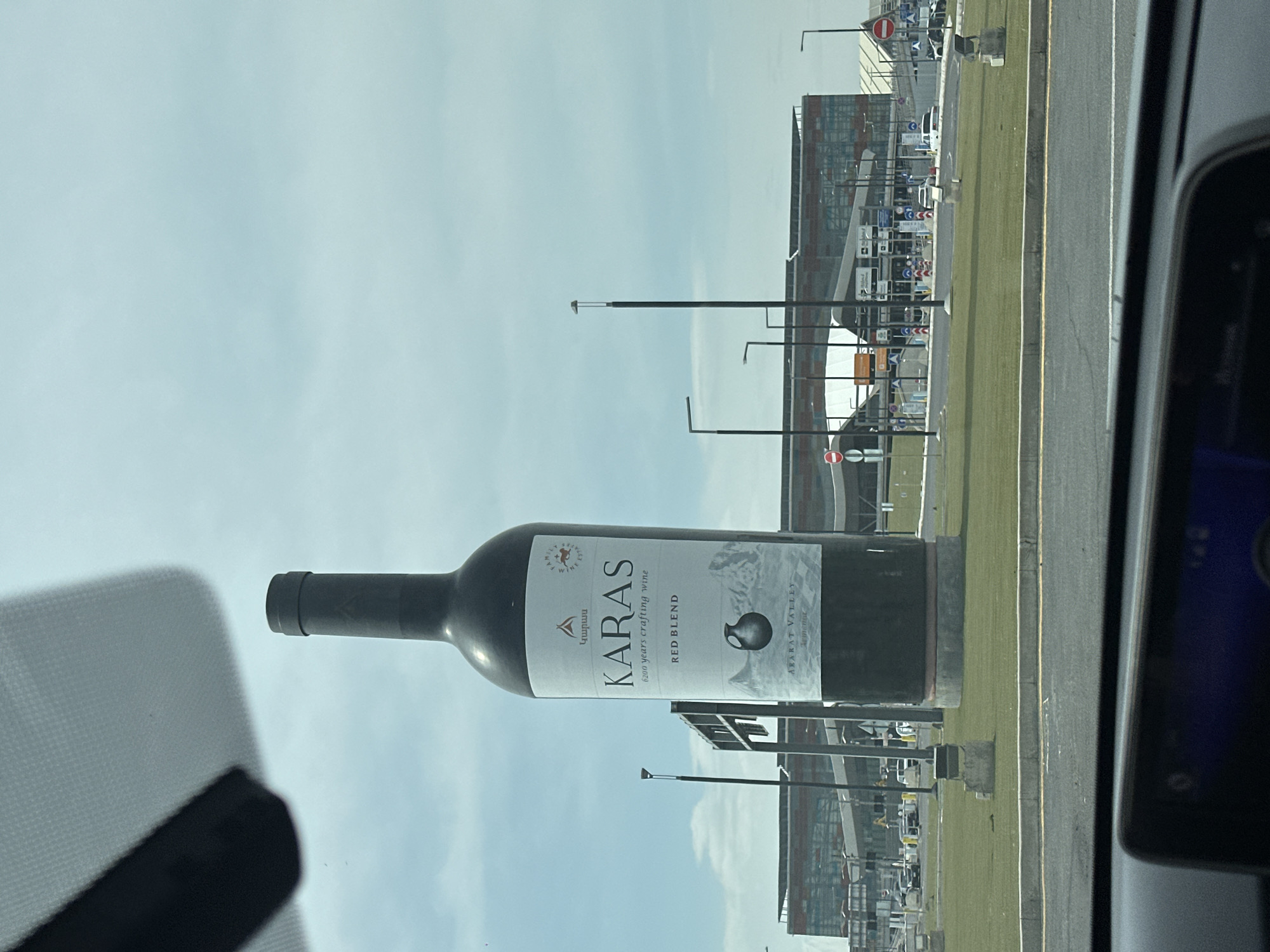 The image shows a large, billboard-sized model of a bottle of Karas red blend wine. The bottle is oriented horizontally and displays the brand name and label prominently. In the background, there is an airport terminal-like building and a roadway with light poles. The shot appears to be taken from inside a vehicle, possibly near Zvartnots International Airport, Yerevan, Armenia, based on the architectural style of the airport building and the wine brand being a local Armenian product.