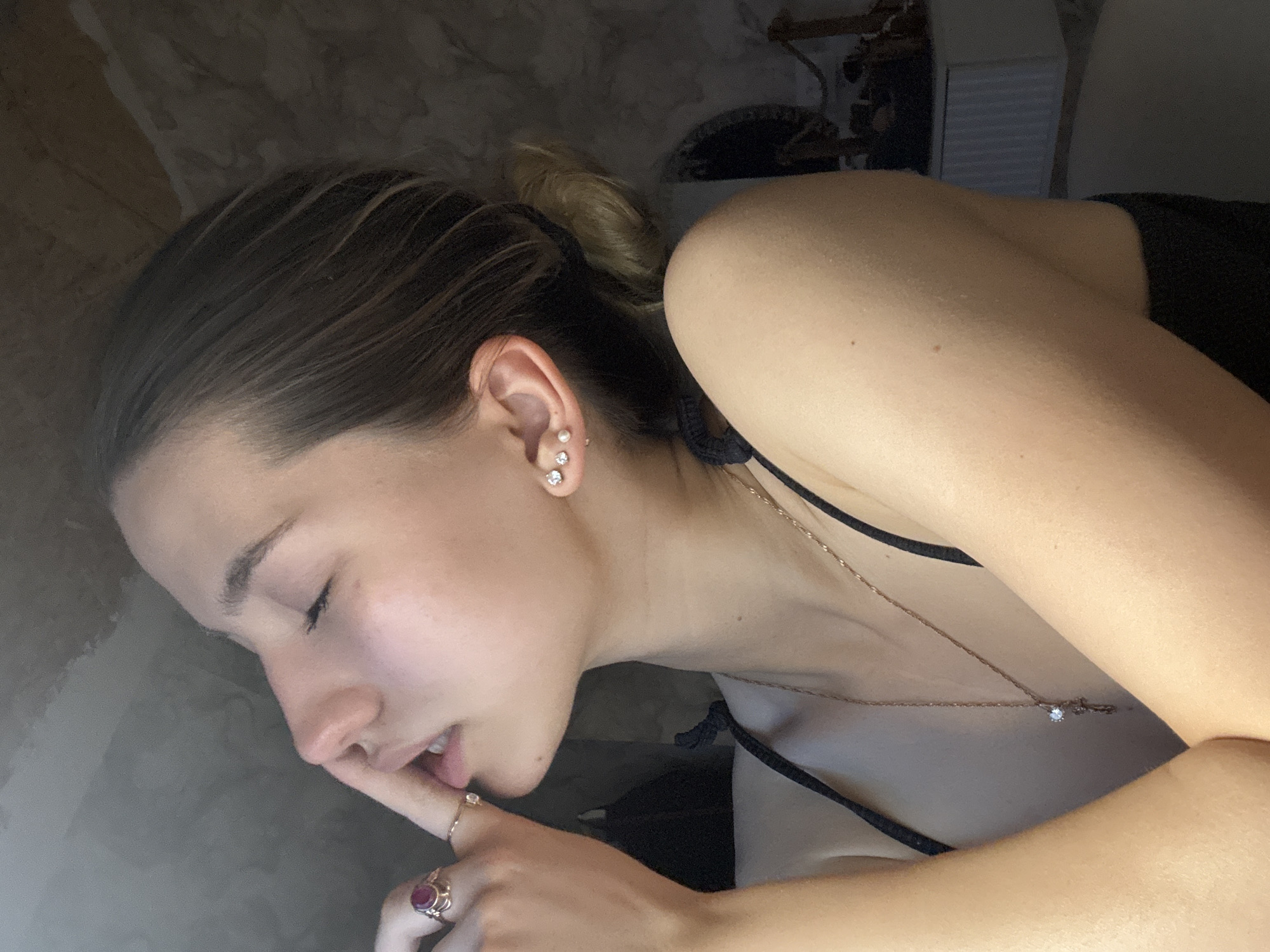 A young woman lies on her side, her head near the top left of the image, and her body extends towards the lower right. Her hair is pulled back in a loose bun, and she is wearing earrings and a necklace with a diamond pendant. She is wearing a black tank top. She has two rings on her finger. She appears to be resting her finger on her lips. The background is out of focus and appears to be a wall, possibly with a marble pattern. There is additional blurry content behind her shoulder. There is no indication of the city or a specific location in Noviy, Russia.