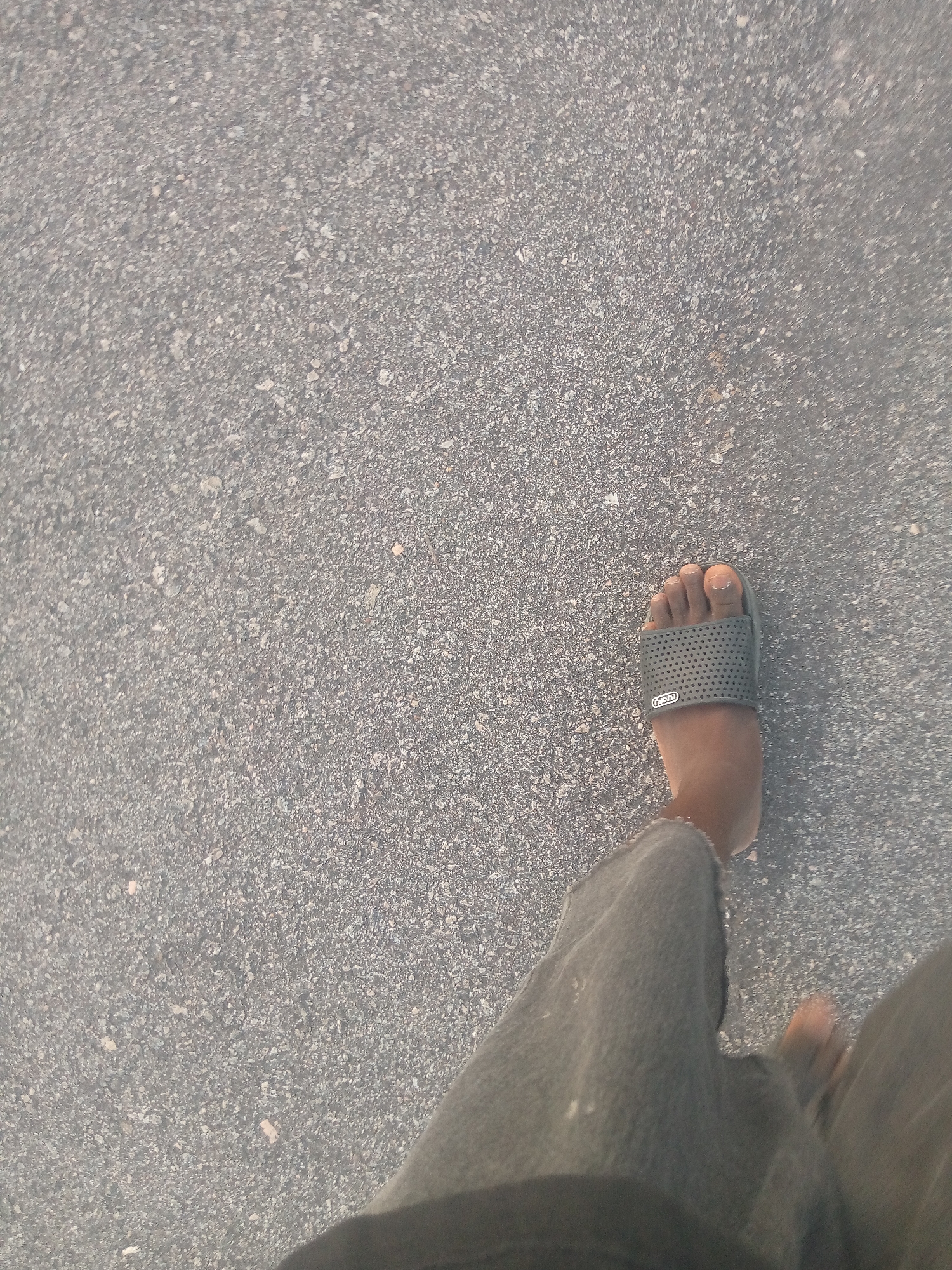 The image shows a close-up, first-person perspective of a person's left foot wearing a dark gray slide sandal with a perforated strap. The foot is stepping on an asphalt surface, which is composed of small, gray aggregate. The background is entirely this asphalt. The person appears to be walking, as indicated by the slight motion blur in the foot and the lower leg, which is clad in dark gray fabric, likely pants. The lighting suggests it is daytime, with sunlight casting a slight shadow. The image was taken in Jauro Yinu, Nigeria.