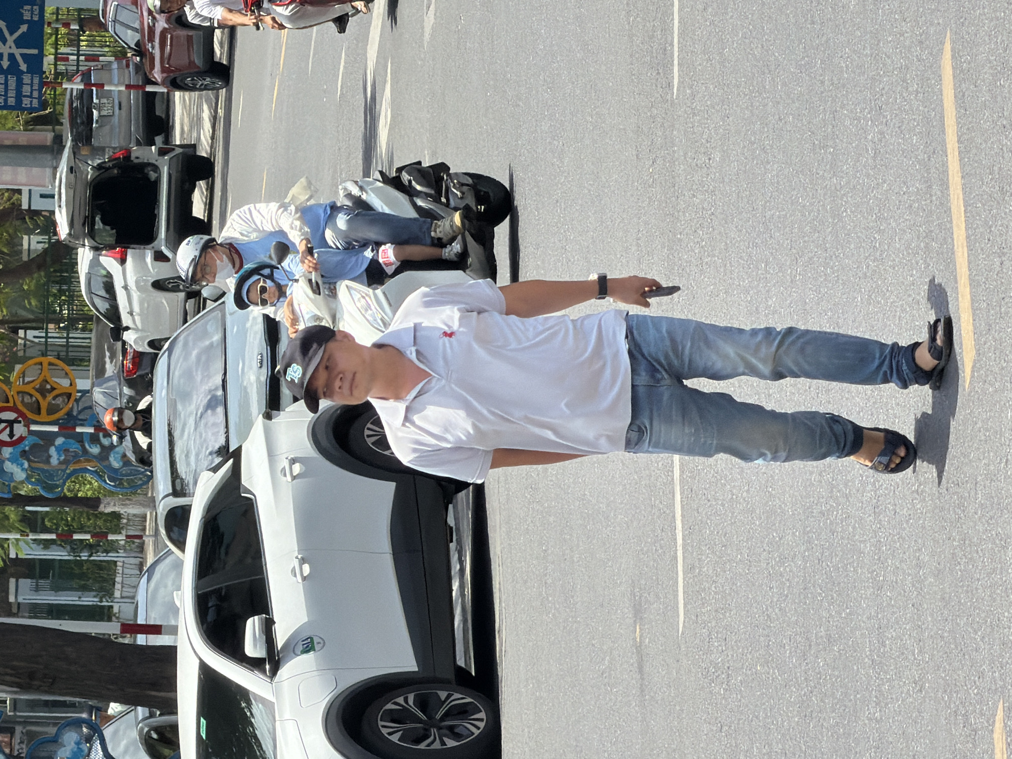 In Nha Trang, Vietnam, a man wearing a light blue polo shirt, blue jeans, and dark sandals is walking across a paved street. He is holding a dark smartphone in his right hand. In the background, a person wearing a white shirt and helmet is riding a scooter with a child in front, also wearing a helmet. Several cars are parked and driving on the street, including a white electric vehicle and a red SUV. The environment appears to be a city street with trees and buildings visible.