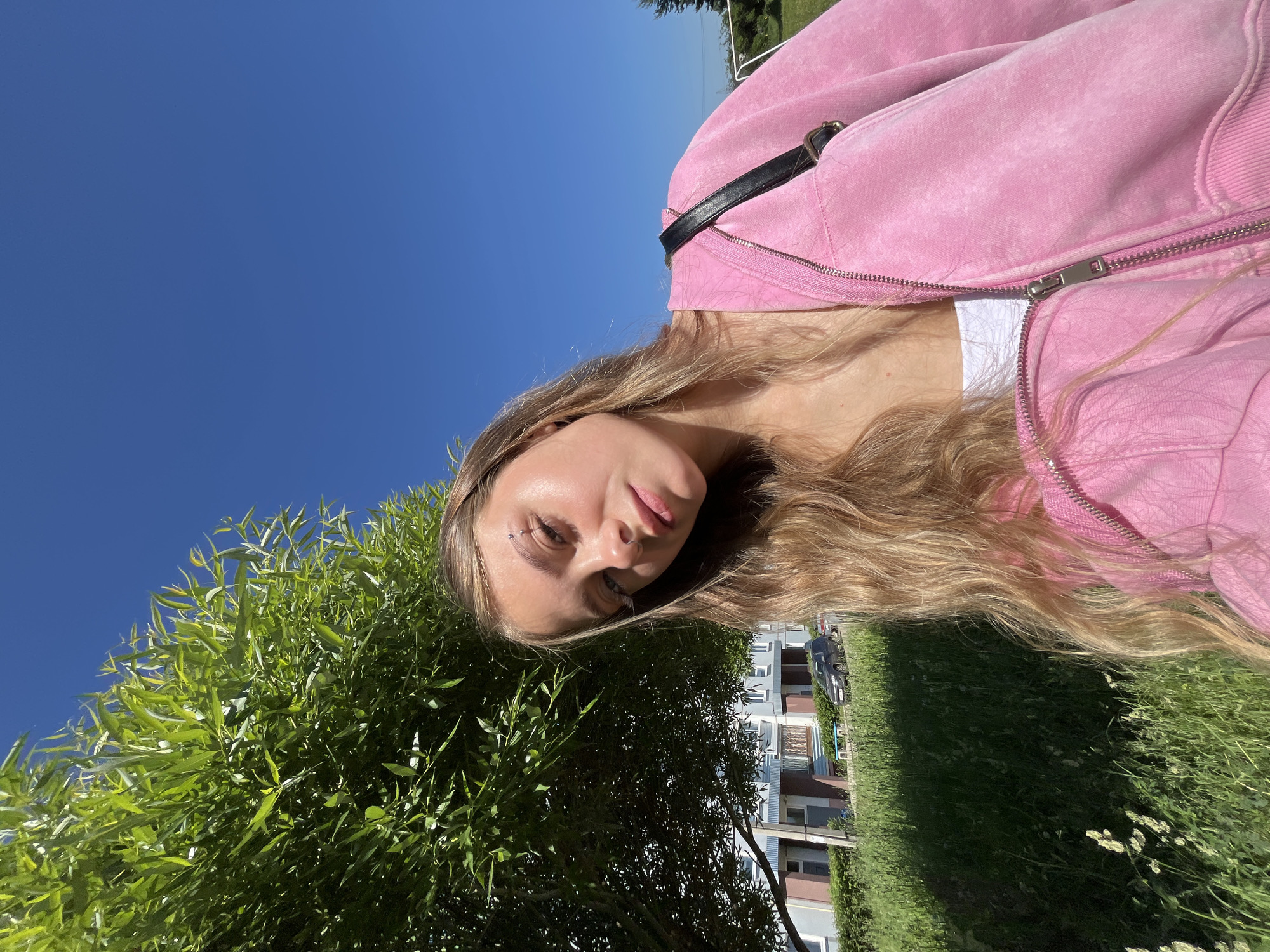 The image shows a young woman with long blonde hair, wearing a pink zip-up hoodie and a white top, outdoors on a sunny day. She has a piercing above her eyebrow and a nose ring. The background includes a large green tree, lush grass, a building with multiple stories, a street with a parked car, and a clear blue sky. The scene appears to be in a residential area.