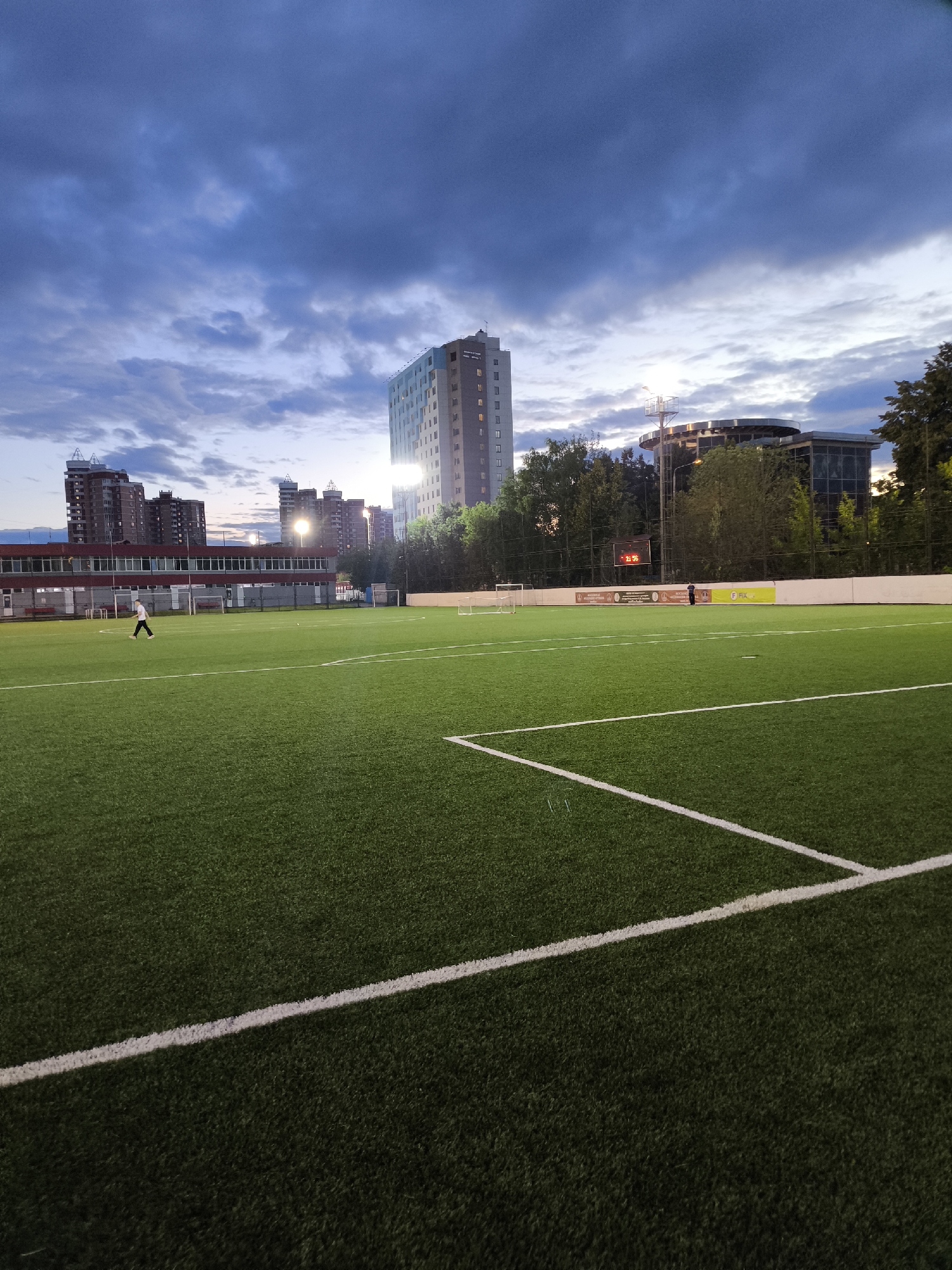 The image depicts an outdoor artificial turf soccer field at dusk. The sky is filled with dramatic clouds, suggesting an overcast or stormy weather condition. In the background, several tall buildings are visible, indicating an urban or suburban setting. On the left side of the field, a person is walking. Further in the background, there is a scoreboard displaying the time "2:56" and some text that is too small to read clearly. A fence runs along the perimeter of the field, with banners visible behind it. The overall atmosphere is calm and slightly moody, possibly due to the twilight setting and the cloudy sky. The presence of the soccer field, goalposts, and markings on the turf confirm the activity taking place.