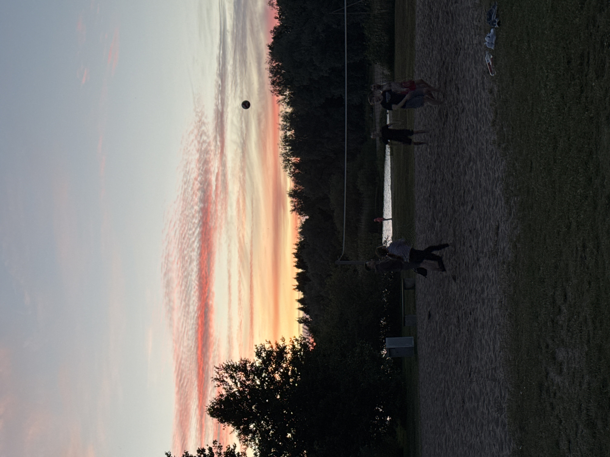 The image shows a group of people playing volleyball on a sandy court during sunset. The sky is filled with dramatic, wispy clouds painted in shades of pink, orange, and white, indicating the end of the day. Tall trees form a dark silhouette against the vibrant sky, bordering the court. A volleyball net is visible, and several individuals are actively engaged in the game, with one person in mid-air, seemingly hitting the ball. The sandy ground suggests a beach or a dedicated sand court, and the presence of grass on the right side of the image indicates a park or recreational area. The overall atmosphere appears energetic and recreational, with the beautiful sunset adding a serene and picturesque quality to the scene. No visible text is present in the image.