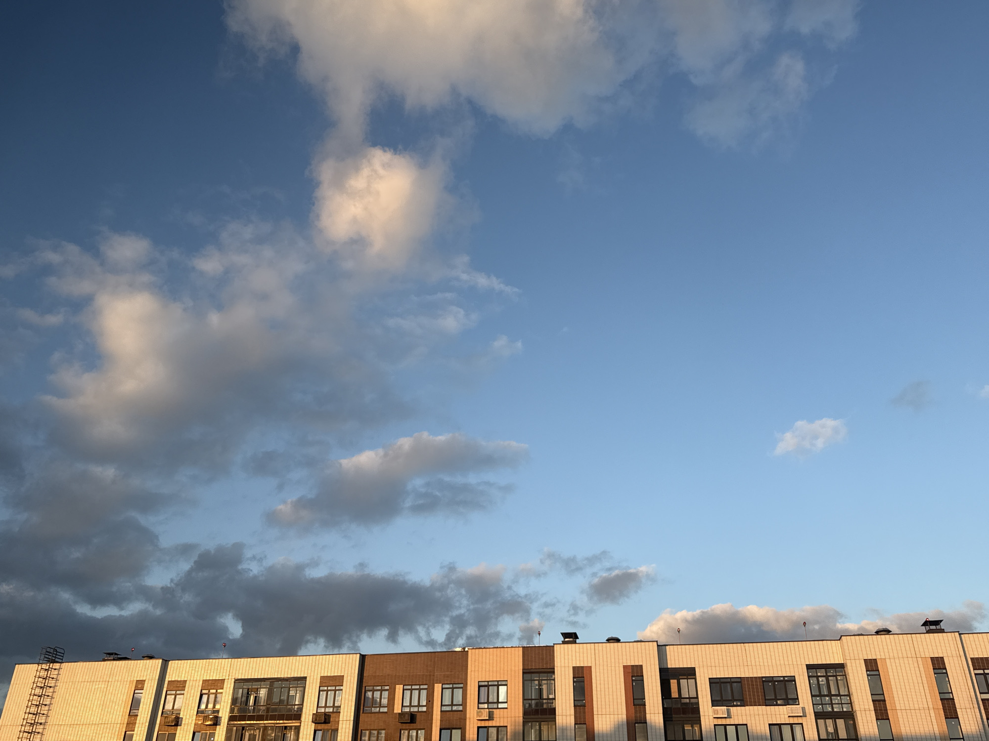 This image captures the upper floors of a modern residential building against a backdrop of a blue sky with scattered clouds. The building features a varied facade with different sections clad in light beige and dark brown tiles. Many windows, some with balconies, are visible, reflecting the sky. On the roof, ventilation pipes and possibly antennas are present. The lighting suggests it is either sunrise or sunset, with warm light illuminating parts of the building and some clouds. The image does not contain any people, actions, or interactions. The scene is likely from Lyubertsy, Russia, given the context provided.