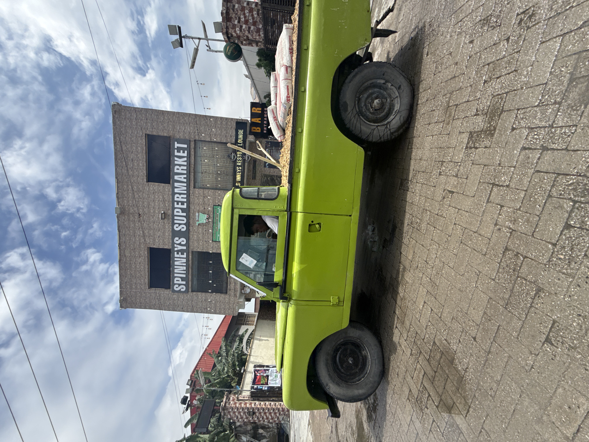 A lime green pickup truck is parked on a wet, patterned brick road in front of a building. The building has a sign that reads "SPINNEYS SUPERMARKET". Above the supermarket, there is a sign for "HUNTERS BAR ENTRANCE LOUNGE". The truck bed is filled with several large white bags, possibly cement. There are also some dry goods like grains or seeds in the truck bed. A person is visible inside the truck's cab. The sky is partly cloudy, suggesting it is daytime. The overall scene appears to be a commercial area in Ikorodu, Nigeria.