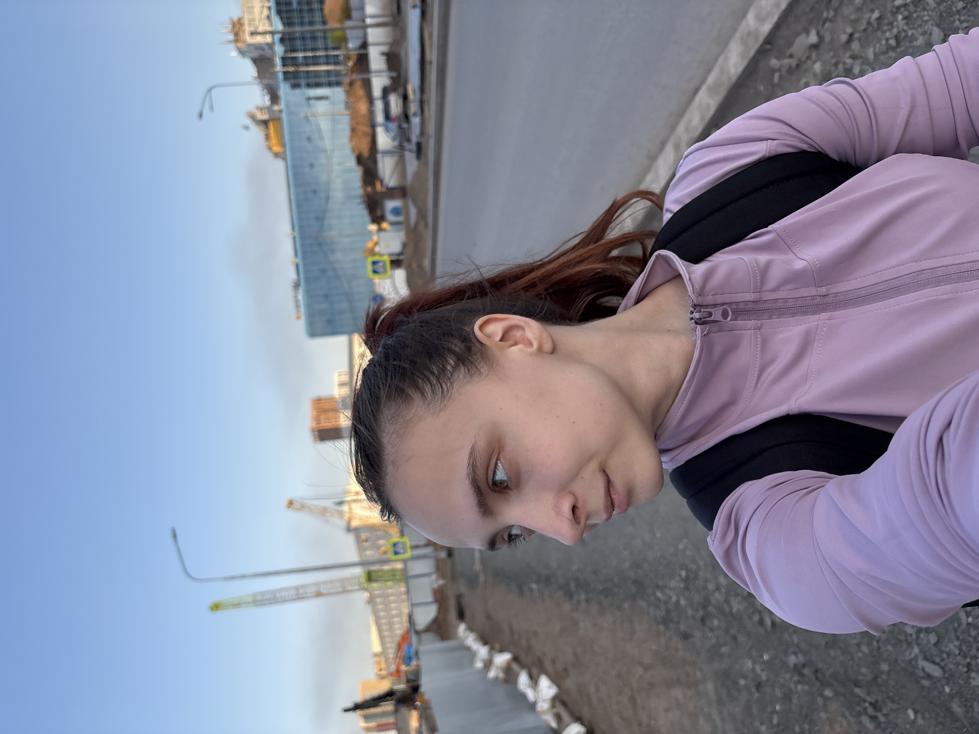 A young woman is outdoors, likely in Perm, Russia, identifiable by the presence of construction cranes and buildings typical of urban development in the background. She is wearing a light purple zip-up jacket with a black backpack. The setting appears to be alongside a road or walkway under construction, indicated by the uneven ground, construction materials, and partially completed infrastructure. The sky is clear and suggests either early morning or late afternoon lighting.