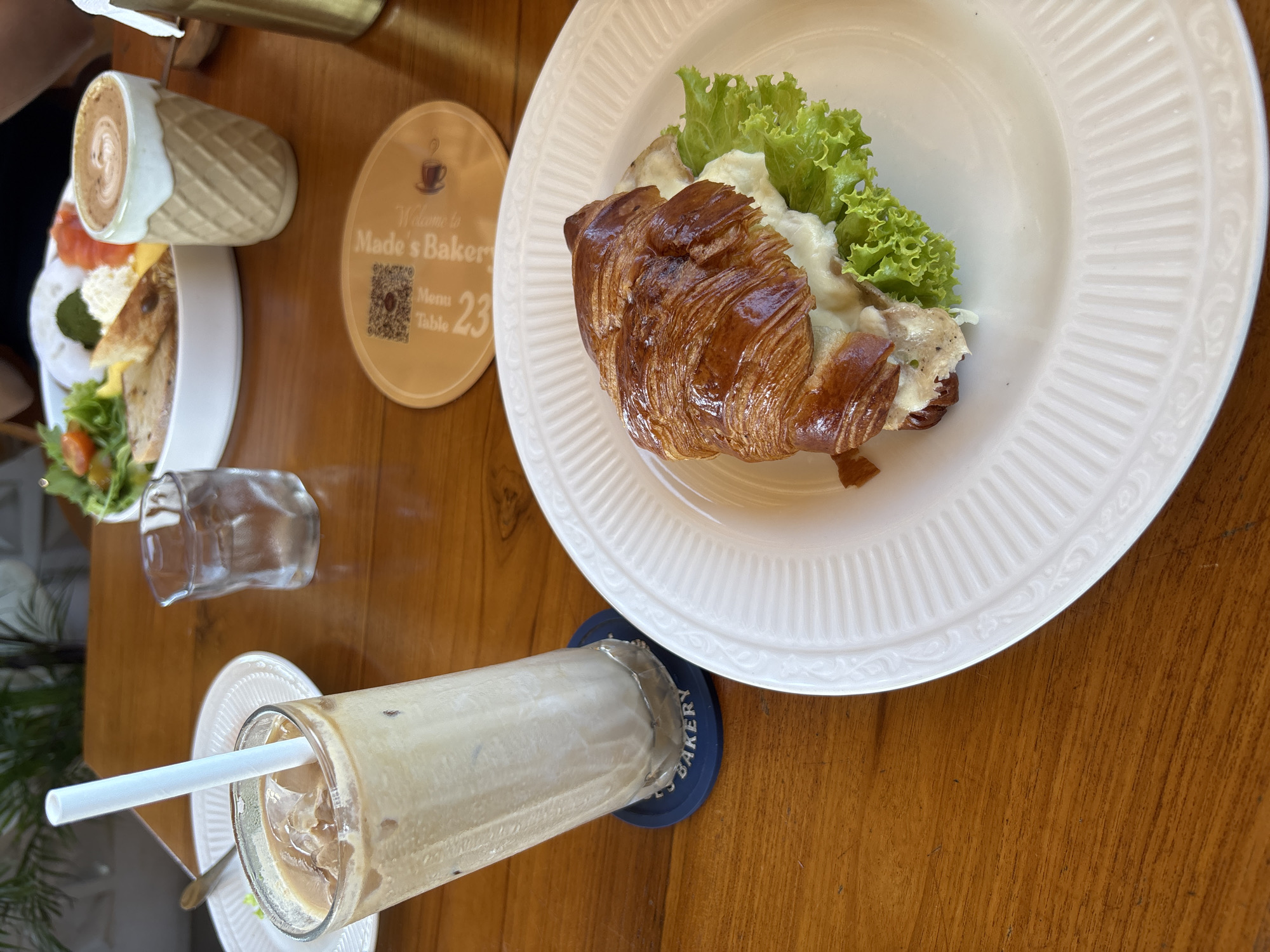 This image shows a table setting at Made's Bakery in Kuta Selatan, Indonesia. A plate with a croissant sandwich filled with lettuce and what appears to be cheese sits on a white plate. A tall glass of iced coffee with a straw rests on a blue coaster with the bakery's name. Nearby is a smaller dish with a latte and salmon, and a small glass of water. A yellow coaster with the bakery's name and table number (23) is also visible. The table itself is wooden. No people are in focus.