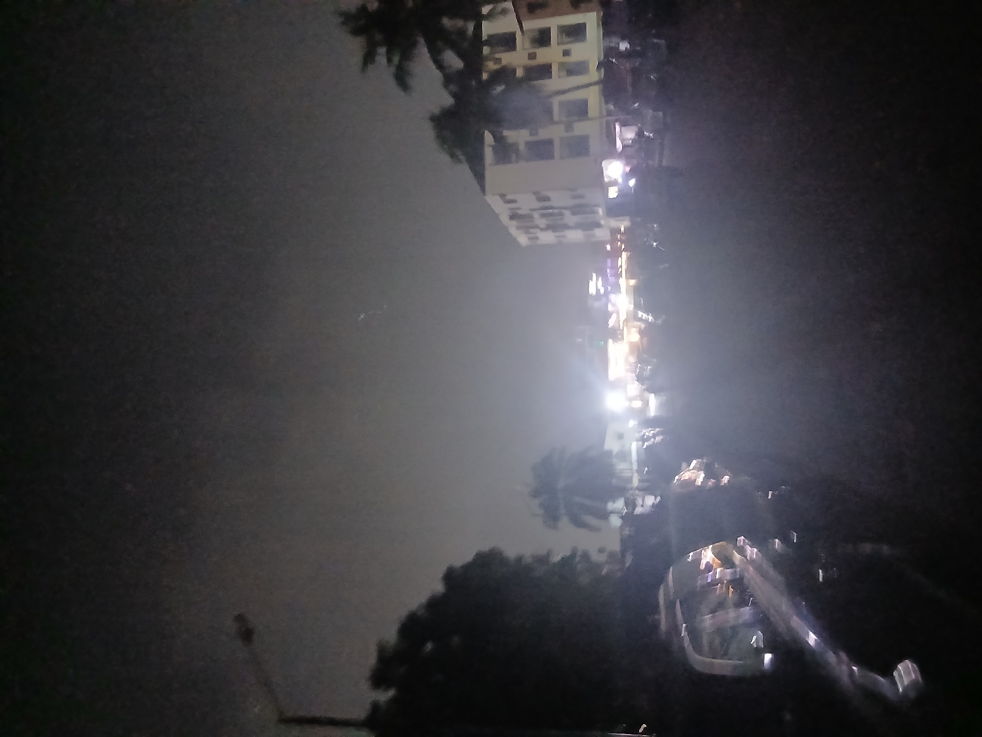 The image shows a poorly lit street in Lagos, Nigeria at night. A multistory building is visible in the upper portion of the image, with streetlights illuminating a section of the road. A palm tree is silhouetted against the sky near the building. A vehicle with a visible side mirror is in the foreground, and the surrounding area is dark. There are no identifiable people present.