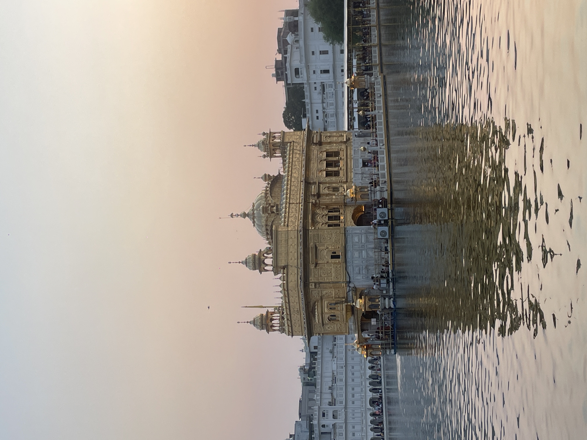 The image shows the Golden Temple in Amritsar, India. It is a Sikh gurdwara, and is considered the holiest site in Sikhism. The temple is made of gold and is surrounded by a large pool of water. The temple is illuminated by the soft light of the setting sun. There are people on the walkway surrounding the temple, and some are in boats on the water. The sky is a gradient of pale orange and pink, with some faint clouds. The water reflects the golden colors of the temple and the sky. The buildings in the background are white with arches.