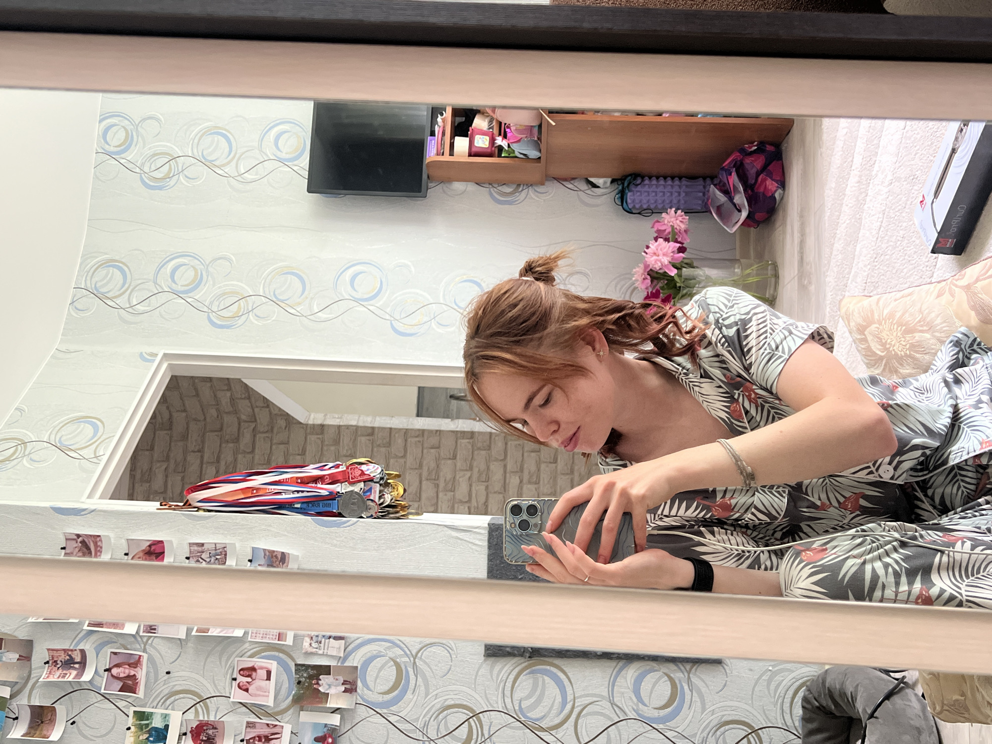 A young woman with auburn hair tied into two ponytails is taking a selfie in a mirror. She is wearing a grey pajama set with a tropical leaf and flamingo print. Her hair is styled in braids that fall over her shoulders. She is wearing a silver ring on her left ring finger and a black watch on her left wrist. In the reflection, a wall with framed pictures and medals is visible. There is also a door frame and a brick wall in the background. To the right of the mirror, a wooden shelf unit with various items is visible, including a television, a vase of pink flowers, and a purple foam roller. A patterned rug covers the floor. The scene appears to be an indoor setting, likely a bedroom or living area, in Chaikovsky, Russia.
