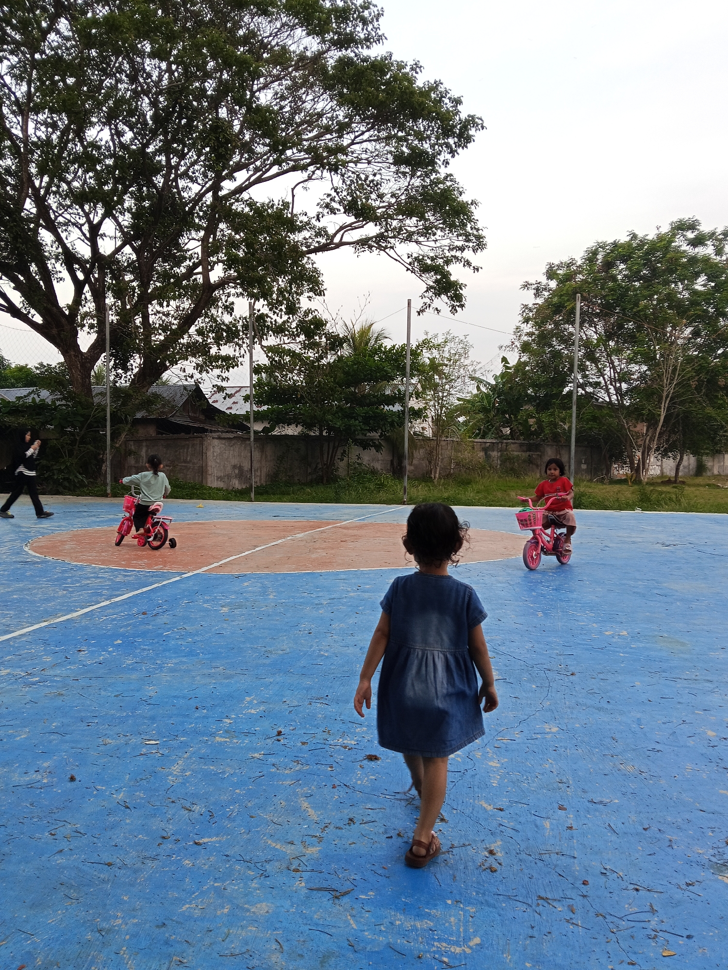 The image depicts an outdoor scene on a sports court in Lhokseumawe, Indonesia, featuring four individuals. In the immediate foreground, a young child, seen from behind, walks away from the camera. This child wears a blue denim dress and sandals, and has dark hair. The court surface is predominantly blue with worn patches, white lines, and a large orange semi-circle, suggesting a basketball or badminton court. Debris like small sticks and leaves are scattered across the blue surface.
Further back on the court, two other young children are riding small pink bicycles equipped with training wheels. One child, wearing a light green top, is on the left side of the frame, riding towards the left. The other child, dressed in a red top, is on the right, riding towards the center.
In the mid-ground, an adult woman, dressed in a black top, white trousers, and a head covering, walks from left to right. The background is characterized by dense foliage from large trees, particularly on the left and right sides of the frame. Fences, a concrete wall, and simple building structures are visible beneath the trees. The sky is overcast and bright, indicating either early morning or late afternoon. The overall atmosphere suggests a casual, playful outdoor activity. No visible text is present in the image.