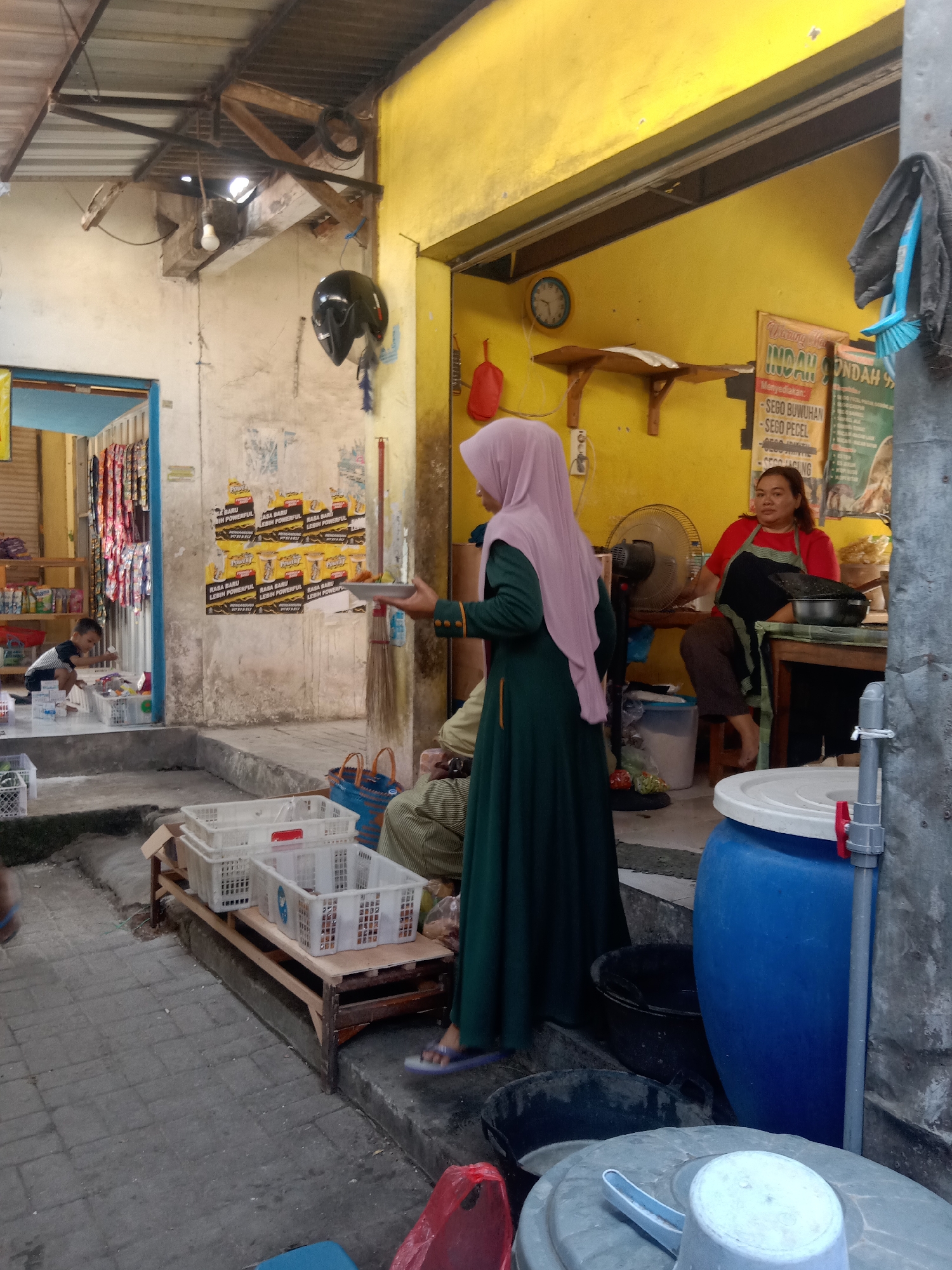 The image depicts a street-level scene, likely a food stall or small eatery, in Klepek, Indonesia. In the foreground, a woman wearing a green dress and a light pink hijab is holding a tray of food. To her right, a woman in a red shirt and a dark apron is seated at a table, appearing to be serving or preparing food. Behind them, a bright yellow wall serves as the backdrop for a menu board listing various Indonesian dishes, such as "SEGO BUWUHAN" and "SEGO PECEL."

The environment is rustic and somewhat cluttered. To the left, a child in a dark shirt and shorts is seen kneeling near some white plastic baskets filled with items. Shelves stocked with goods are visible in the background, suggesting a combined shop or storage area. Various containers, including large blue and gray plastic barrels, are present, along with a fan and a helmet hanging on the wall. The overall setting appears to be an open-air market or a roadside establishment, with visible structural elements like wooden beams and a corrugated metal roof. The ground is paved with stone tiles.