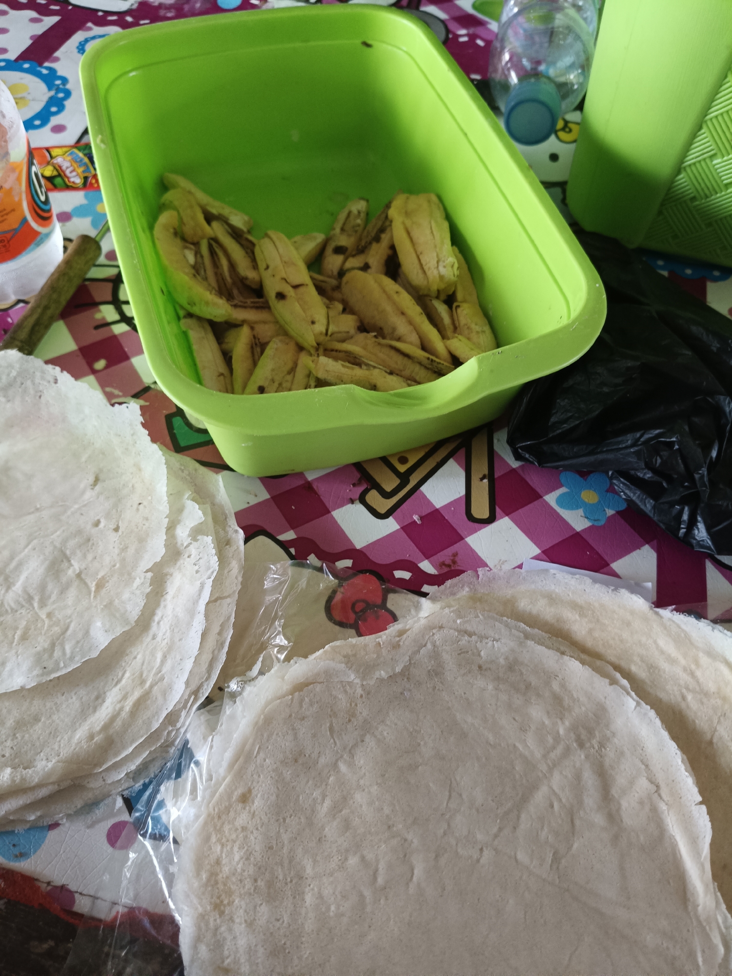 The image depicts a close-up view of food preparation, specifically of what appears to be raw spring roll wrappers and banana slices, likely in a domestic setting in Rancaseneng, Indonesia. In the foreground, a stack of thin, white spring roll wrappers is visible, some of which are separated by clear plastic packaging. To the right, another stack of these wrappers is more clearly discernible. In the background, a bright green plastic container holds numerous slices of peeled banana, some exhibiting browning on their edges, suggesting they may have been recently peeled or are slightly overripe. A black plastic bag lies to the right of the container. The surface beneath the food items is covered by a colorful, checkered tablecloth with a pattern of pink, white, and some blue and yellow flowers. A clear plastic bottle with a blue cap is also visible behind the green container. The overall scene suggests a home kitchen or dining area where food is being prepared for consumption or cooking. There are no people visible in the image.