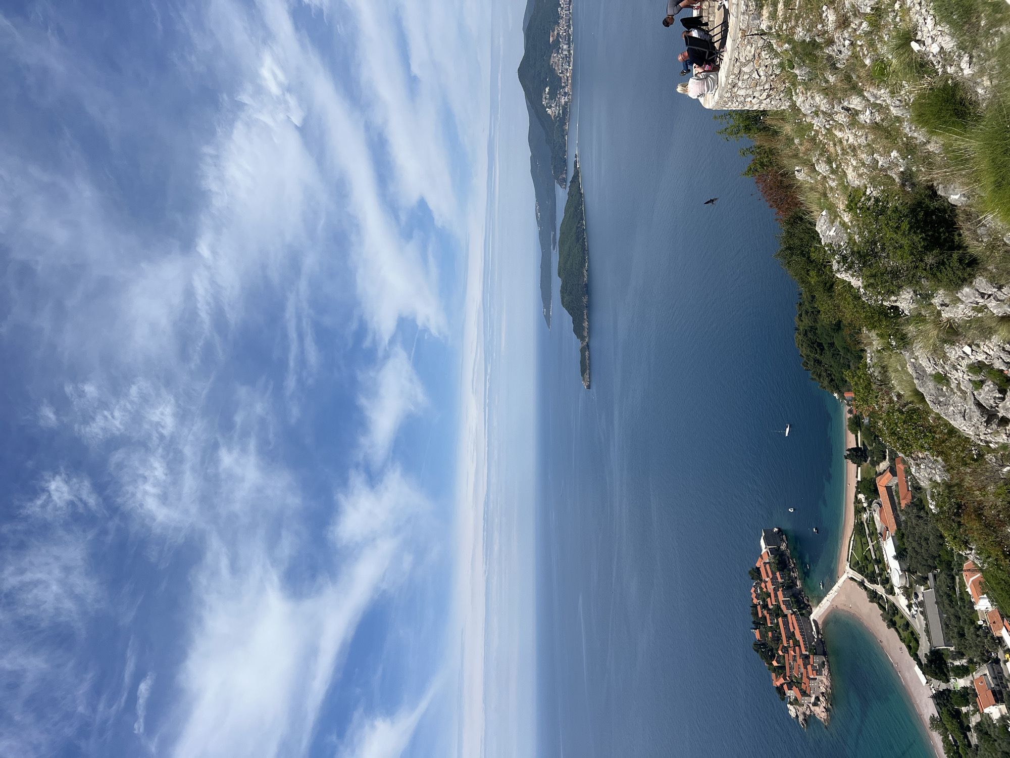 From a high vantage point on a cliff, a view overlooks Sveti Stefan, Montenegro, and the Adriatic Sea. The sky is blue with wispy clouds. The coastline features a distinctive islet village with terracotta rooftops connected to the mainland by a sandy isthmus. Buildings line the beach. Further out, elongated islands are visible in the sea, fading into the horizon. In the upper right corner, a few people are sitting along the cliff. A bird is flying near the cliffside.