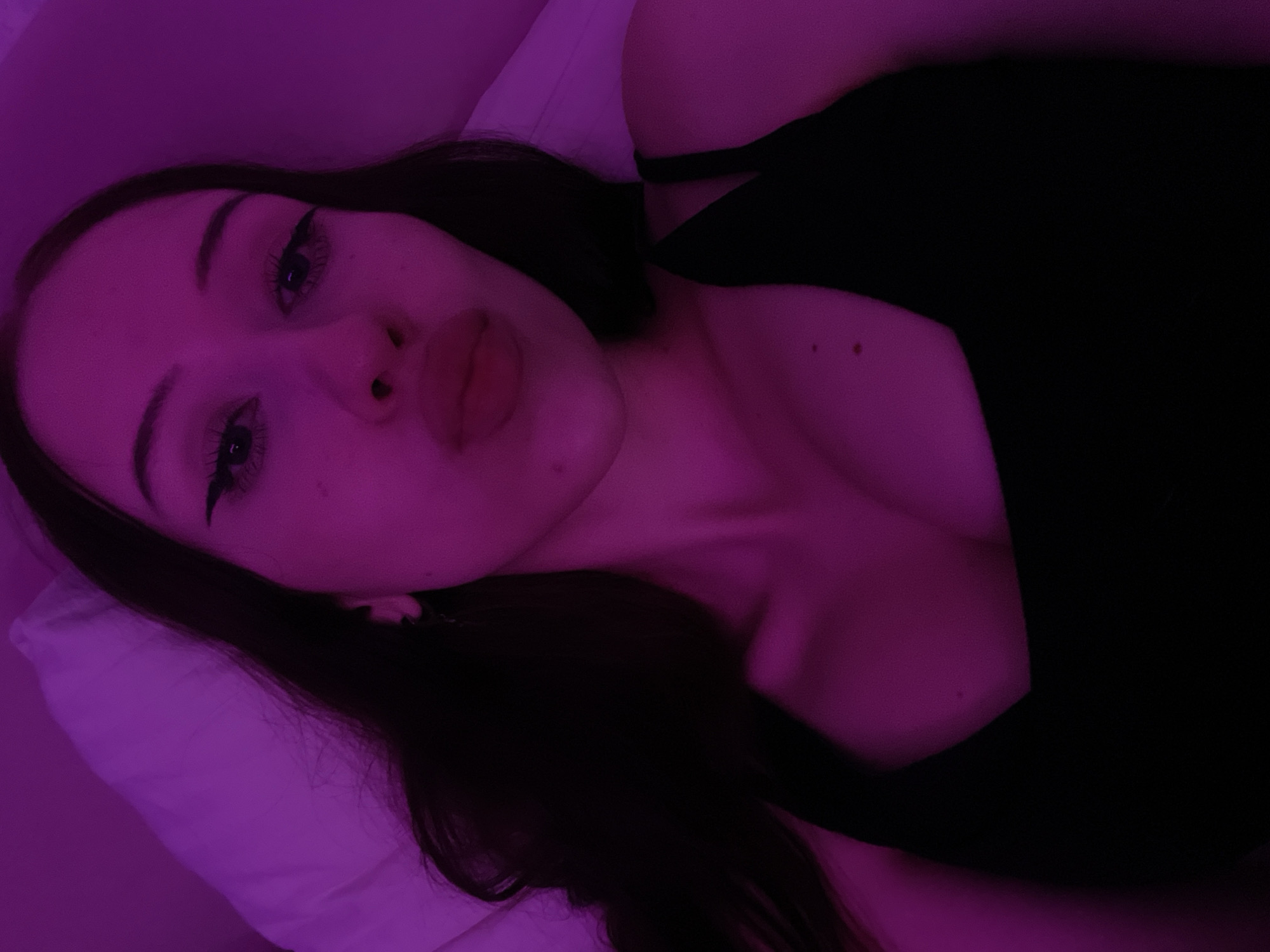 The image shows a close-up, selfie-style shot of a young woman lying down. She has long, dark hair and is wearing a black top. Her face is illuminated by purple light, giving her skin a pinkish hue. Her lips are slightly parted, and she has noticeable eyeliner. The background appears to be a pillow and a wall, also lit by the purple light. There is no discernible information in the image to indicate a location in Votkinsk, Russia.