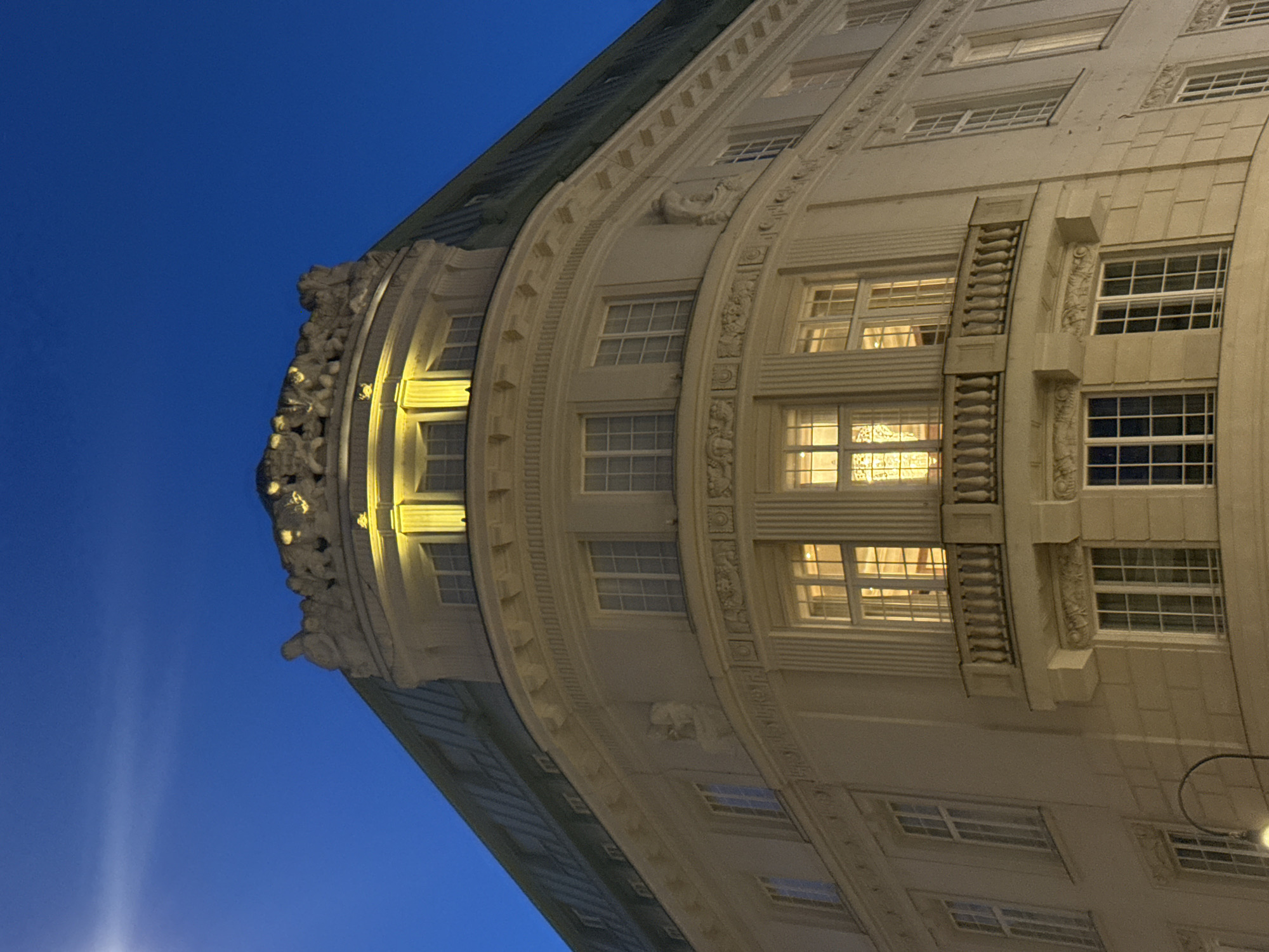 A detailed, forensic, matter-of-fact description of the scene from Vienna, Austria, is as follows:
The image is an upward-facing, medium-shot captured at twilight of the ornate facade of a multi-story building, likely an apartment or office building, situated on a street corner in Vienna, Austria. The sky above is a deep, clear twilight blue with a hint of lighter blue near the horizon.
The building itself is constructed of a light-colored stone, possibly sandstone, with classical architectural elements. It features a curved corner, accentuating its grandeur. The roofline is adorned with a decorative frieze depicting a series of sculpted figures, possibly mythological or allegorical in nature, illuminated by warm, yellow lights positioned along the top of the facade. These lights cast a glow downwards, highlighting the architectural details of the upper floors.
The building has multiple levels of windows. Many of the windows on the upper floors are lit from within, emitting a warm, inviting yellow light, suggesting occupancy. These windows are rectangular and appear to have multiple panes. One prominent feature is a balcony on the second story of the curved corner, with a balustrade made of stone or concrete, featuring a repeating decorative pattern. Below the balcony, there is another decorative band above a row of windows.
Further down, on the lower levels, some windows are dark, while others show signs of interior lighting. The facade is detailed with cornices, moldings, and recessed panels. A street lamp with a curved arm and a visible light fixture is partially visible in the lower left corner of the image, suggesting the scene is at street level or slightly above. The building's overall style suggests it is from the late 19th or early 20th century, consistent with Viennese architecture of that period. No people are visible in the image.