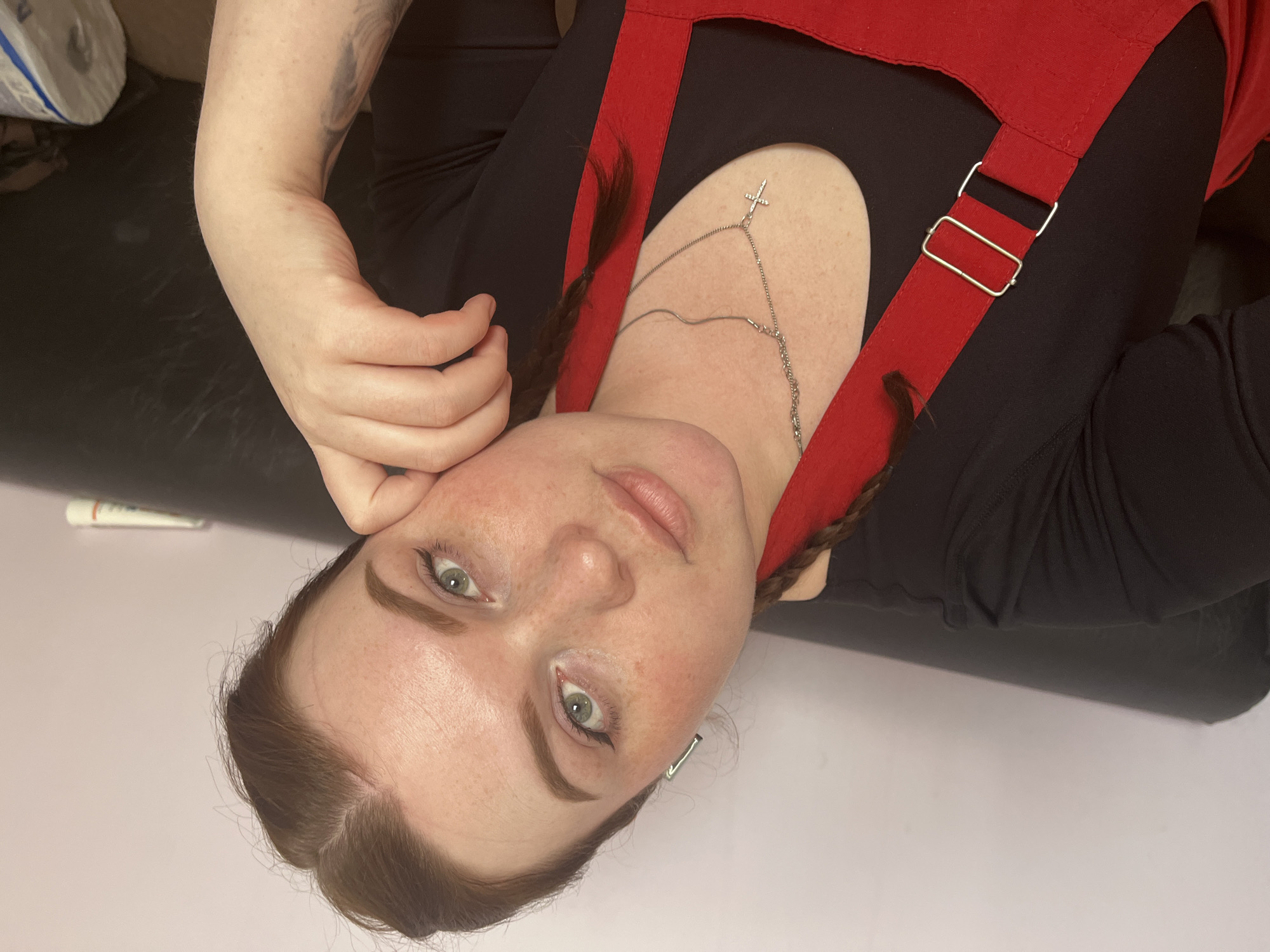 A fair-skinned woman with green eyes is lying on a surface. She is wearing a black long-sleeved top and a red garment with straps. A silver body chain with a cross pendant adorns her neck. Her hair is styled in pigtails. A partially visible tube of white and green material is in the background, along with a roll of blue and white material and a black surface. The location is not discernable from the given image.