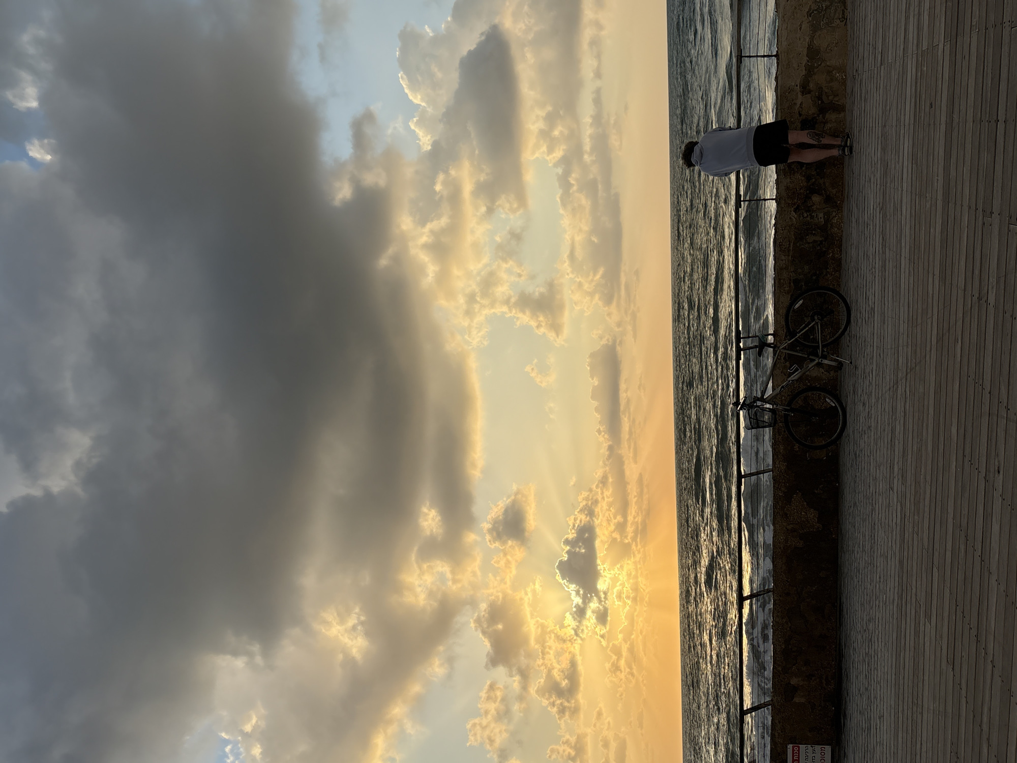 The image captures a picturesque coastal scene at what appears to be sunset in Tel-Aviv, Israel, viewed from a wooden boardwalk or pier. The sky dominates the upper portion of the frame, showcasing a dramatic interplay of clouds. On the left, the clouds are dark and heavy, while towards the right, they are illuminated by intense golden light from the setting sun, with distinct crepuscular rays piercing through.
In the foreground, a person stands at the edge of the wooden boardwalk, looking out towards the sea. The person is seen from behind, wearing a light-colored top and dark shorts, appearing to contemplate the expansive view. A dark-colored bicycle with a front basket is parked beside them, leaning against a low stone wall or concrete barrier that separates the boardwalk from the water.
The sea stretches across the mid-ground, with gentle waves reflecting the warm, golden hues of the sky, creating shimmering patterns on the water's surface. A metal railing runs along the top of the barrier, partially obscured. The overall atmosphere is one of serene beauty, enhanced by the stunning natural light.
On the barrier, near the bottom right corner, a red and white sign with Hebrew text is visible, though too small and indistinct to be legible.