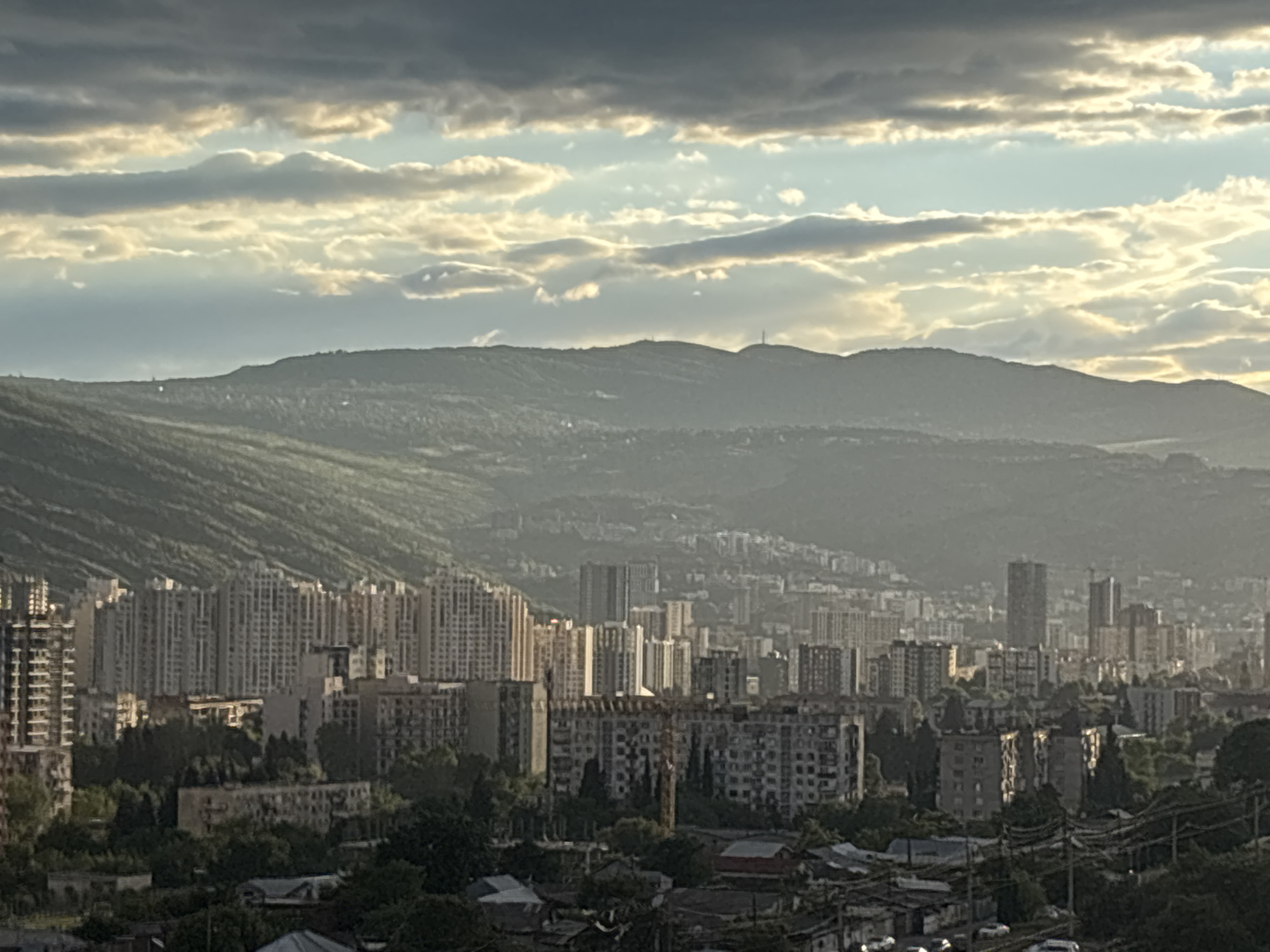 The image shows a panoramic view of Tbilisi, Georgia, with a hazy atmosphere. In the foreground are low-rise residential buildings and dense foliage. Further back, a dense urban landscape unfolds, characterized by a mix of Soviet-era apartment blocks and more modern high-rise buildings. Rolling hills serve as a backdrop to the city, partially obscured by atmospheric haze. A radio tower is visible atop the hills. The sky is overcast with textured clouds tinged with sunlight, indicating either dawn or dusk. No people are visible in the shot.