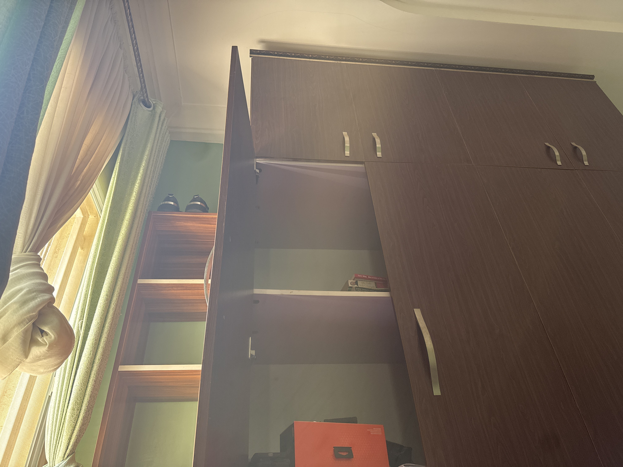 The image shows an interior scene with a large, dark wood wardrobe. One door of the wardrobe is ajar, revealing empty shelves inside. The wardrobe is fitted with silver-toned handles. To the left of the wardrobe, a window is partially covered by draped curtains in shades of cream and pale green. Sunlight filters through the curtains, casting soft light and shadows on the scene.
On top of a shelf unit to the left of the wardrobe, two black and silver shoes are visible. The shelf unit itself is made of wood and appears to have at least three shelves. The wall behind the shelf unit is painted a light green.
Inside the open wardrobe, on one of the shelves, a small stack of books or pamphlets can be seen. One of the books has visible text, including "My Life" and "Bill Gates". Near the bottom of the open wardrobe, a bright orange box with a handle is placed.
The overall impression is that of a bedroom or dressing area. The lighting suggests it is daytime, possibly morning or afternoon, given the soft, diffused sunlight. The scene is calm and appears to be set in a residential location, as indicated by the presence of personal items like shoes and books. The image does not explicitly show any people.