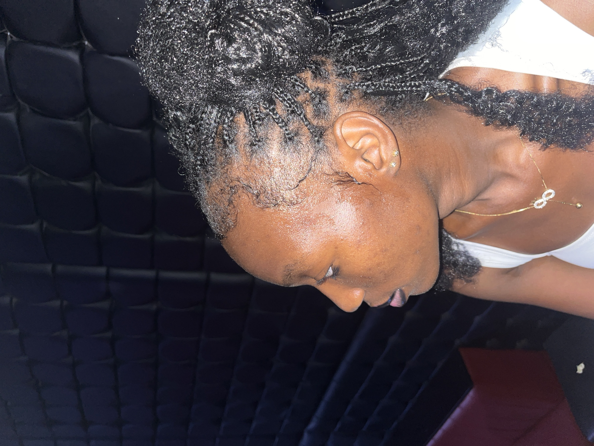 A medium shot shows a Black woman from the side, facing left. She has dark skin, braided hair pulled up, and is wearing a white top and gold jewelry. Her skin appears to be sweating, and she has a small stud earring in her left ear. The background is a dark, quilted wall. A portion of a red and black couch is visible in the bottom left of the frame. The scene is likely indoors, possibly in a club or lounge, and does not provide any specific indicators of location within Howayi, Nigeria.