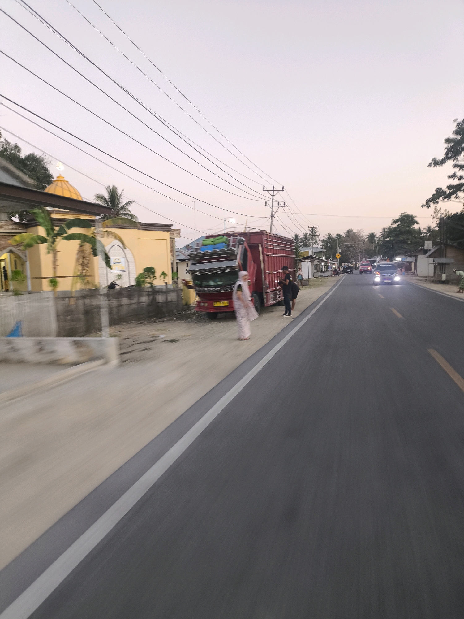 The image captures a scene along a roadside in Dunggala, Indonesia, likely during the late afternoon or early evening, given the soft, diffused light. The foreground is dominated by an asphalt road with a clear white lane marker. To the left of the road, a beige-colored building with a yellow dome and arched window features is visible, possibly a mosque or a religious building. A red truck with a tall, stacked cargo is parked on the side of the road, partially obstructing the view of the building. Two individuals, one wearing a light-colored patterned outfit and the other in darker clothing, are standing near the truck. Further down the road, several cars are driving, with headlights illuminated on one vehicle, indicating diminishing daylight. The background consists of trees, smaller buildings, and utility poles with a network of wires crisscrossing the sky. The overall impression is a typical roadside view in a community, with elements of local architecture, transportation, and daily life.