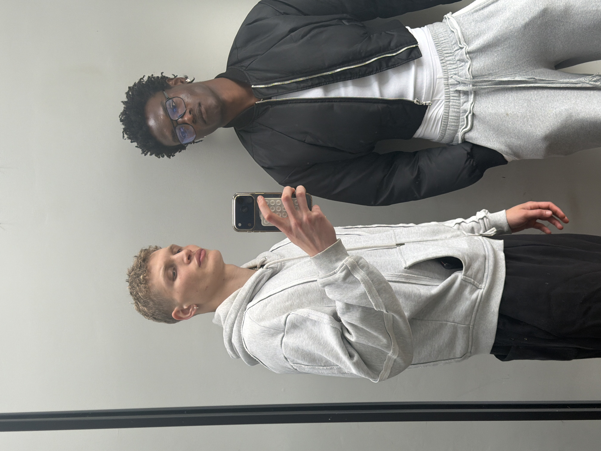 Two males are captured in a mirror selfie against a plain, light-colored wall in what appears to be an indoor setting in London, United Kingdom.
In the foreground, on the left side of the frame, stands a male with light skin and short, light-colored curly hair. He is wearing a light grey full-zip hoodie over a dark top and black trousers. He holds a smartphone with a gold-toned case and a white circular grip or pop-socket, angled as if taking a picture or video. His gaze is directed slightly upwards and to the side, not directly at the phone screen.
Behind him and positioned slightly to the right, is another male with dark skin and dark, curly hair. He wears clear-framed glasses that reflect some light, a black bomber jacket unzipped to reveal a white ribbed top, and light grey sweatpants with prominent elasticized details and drawstrings. He looks directly towards the camera with a neutral expression.
The background is a simple, light-colored wall, possibly off-white or light grey. A dark, horizontal bar or railing is visible across the bottom of the image, suggesting they are standing in front of a large mirror or a window with a reflective quality. The lighting is bright and even, consistent with an indoor environment. There is no visible text in the image. The time of day and weather are not discernible.