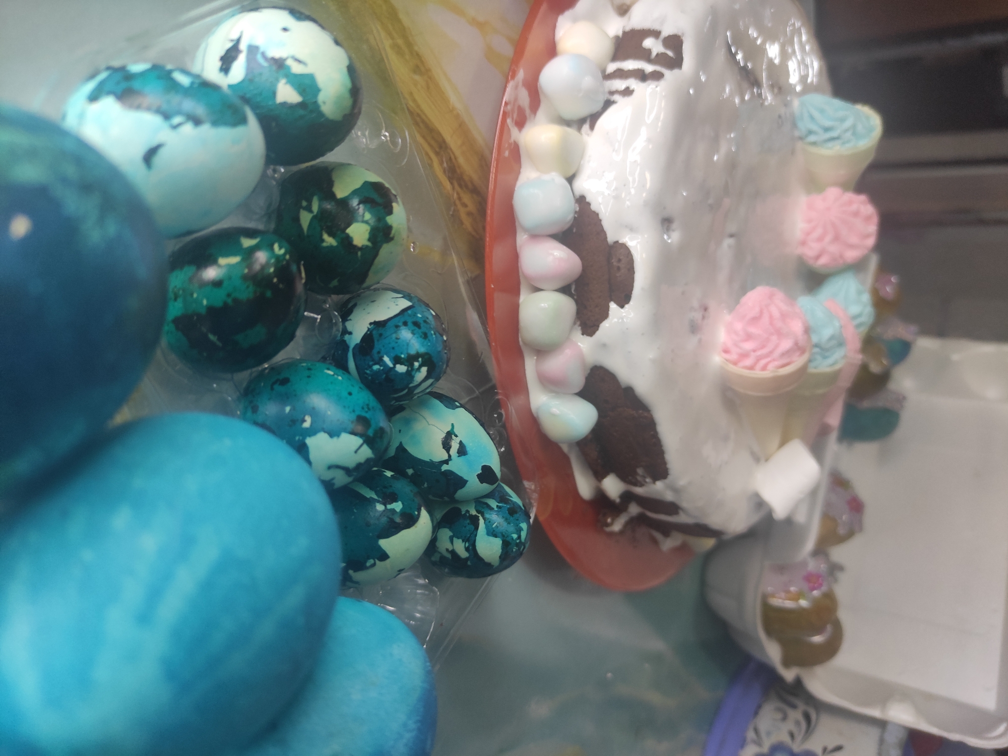 The image shows a close-up of Easter treats, likely photographed indoors. On the left, there's a container of dyed Easter eggs in shades of blue and green, some with a marbled or mottled appearance. To the right, a decorated Easter cake sits on a red plate. The cake appears to be layered chocolate cake with white frosting and is decorated with multicolored marshmallow candies around the edge and miniature ice cream cone decorations. The location is possibly Chaikovsky, Russia, implied by the image metadata, but no visual elements confirm this.