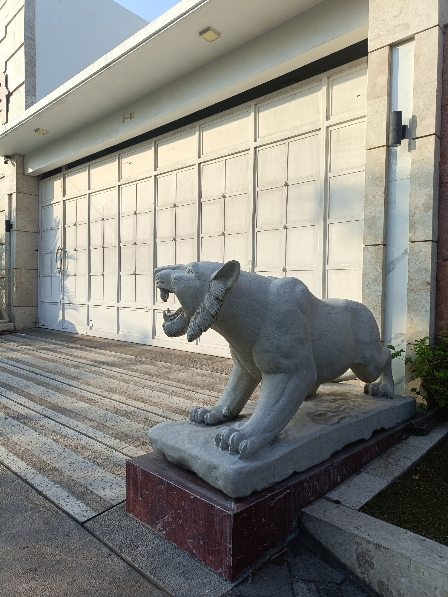 A stone sculpture of a lion in a crouched position is situated on a multi-layered stone platform. The lion sculpture is positioned in front of a large, white, paneled garage door. The area in front of the door is paved with a patterned stone surface, featuring alternating light and dark rectangular tiles. To the right of the garage door, a wall with a dark, cylindrical light fixture attached is visible. A small bush with green leaves is growing to the right of the lion sculpture. The overall scene appears to be an exterior view of a building, possibly a residential or commercial property. The presence of the building and the paving suggests an urban or suburban environment. This image does not provide any specific information that would allow for its location to be identified as Kediri City, Indonesia.