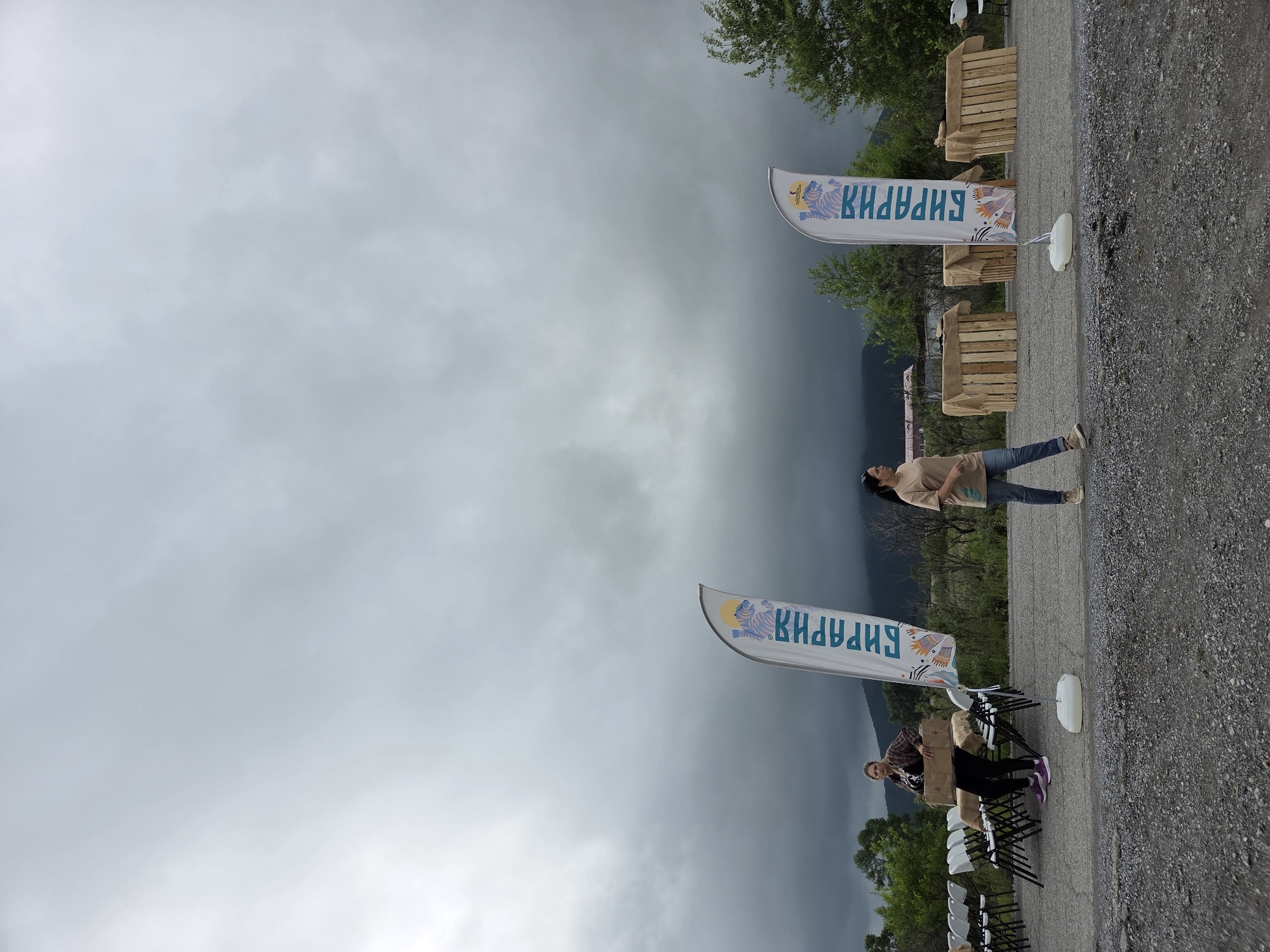 In an outdoor setting, possibly in the city of Teployozersk, Russia, a cloudy sky hangs over a gravelly area. In the foreground, two women are present. The woman on the left, wearing a plaid shirt and dark pants, is carrying a cardboard box. She is standing next to a row of white chairs and tables covered with burlap. A tall, white flag with blue lettering and a graphic of a tiger is planted in the ground near her.
To the right, a woman in a tan t-shirt and jeans is walking. In the middle ground, three wooden structures, also covered with burlap, are arranged. Behind these structures, distant trees and hills are visible. The overall impression is of an outdoor event or gathering being set up.
