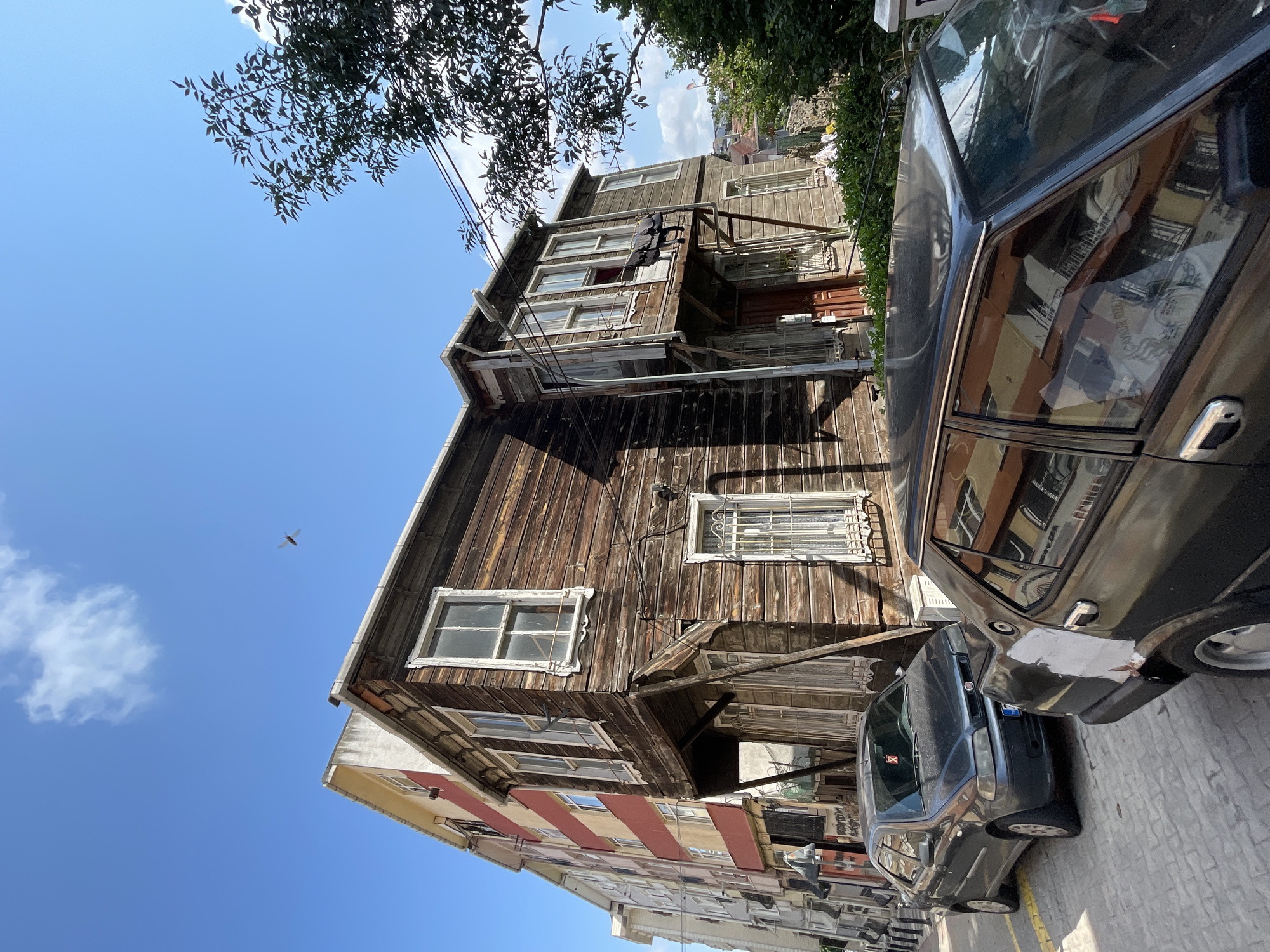 The image depicts a street scene in Kadıköy, Turkey, under a clear blue sky with a few wispy clouds. A vintage wooden house, characterized by its weathered horizontal planks and traditional window frames, stands prominently on the right side of the frame. Adjacent to it, on the left, is a more modern, multi-story building with alternating red and yellow horizontal stripes on its facade.
The street is paved with cobblestones. Two cars are parked along the street. The car in the foreground is a dark-colored sedan, partially visible, showing damage and repair work on its rear quarter panel. Behind it, a dark gray sedan is parked, with a red sticker on its windshield.
The wooden house features a balcony on the upper floor, with laundry hanging on it. There are several windows on both floors of the wooden structure, some with decorative white trim and metal security bars. The more modern building has numerous windows, and air conditioning units are visible on its exterior.
A tree with green foliage grows on the right side of the image, partially obscuring the view of other buildings in the background and power lines. A small insect, possibly a dragonfly, is captured mid-flight in the sky above the buildings. The scene appears to be a quiet urban environment on a sunny day.