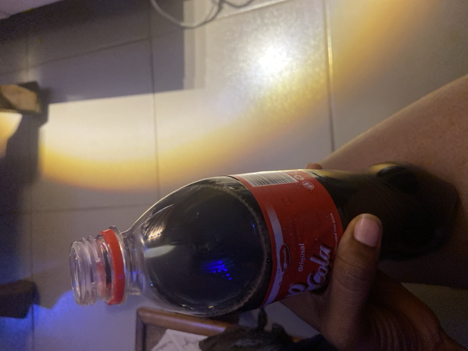 A person holds a plastic bottle of cola in their hand. The bottle is partially filled with a dark liquid. The label is red with white lettering. The background shows a tiled surface with a light reflection creating a gradient. An out-of-focus object is visible below the bottle. The scene is indoors with artificial lighting.