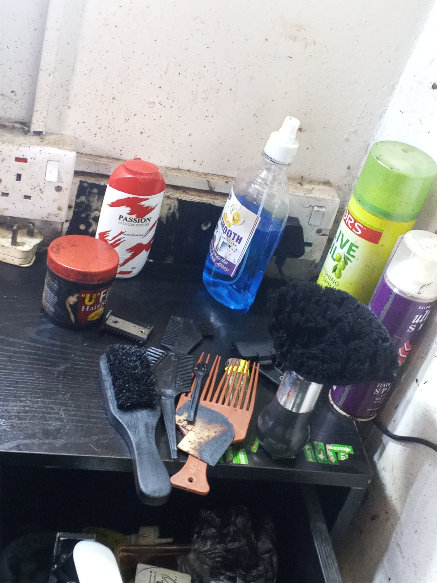 A cluttered shelf area is populated with various hair grooming products. These include, but are not limited to, cans of hair spray, a bottle of shaving lotion, a jar of hair styling product, talcum powder, multiple hair combs, and a brush. The backdrop consists of a wall with electrical outlets. The image seems to be taken in Gwong, Nigeria due to the presence of the product "PASSION Talcum Powder", which is a popular brand in the country.