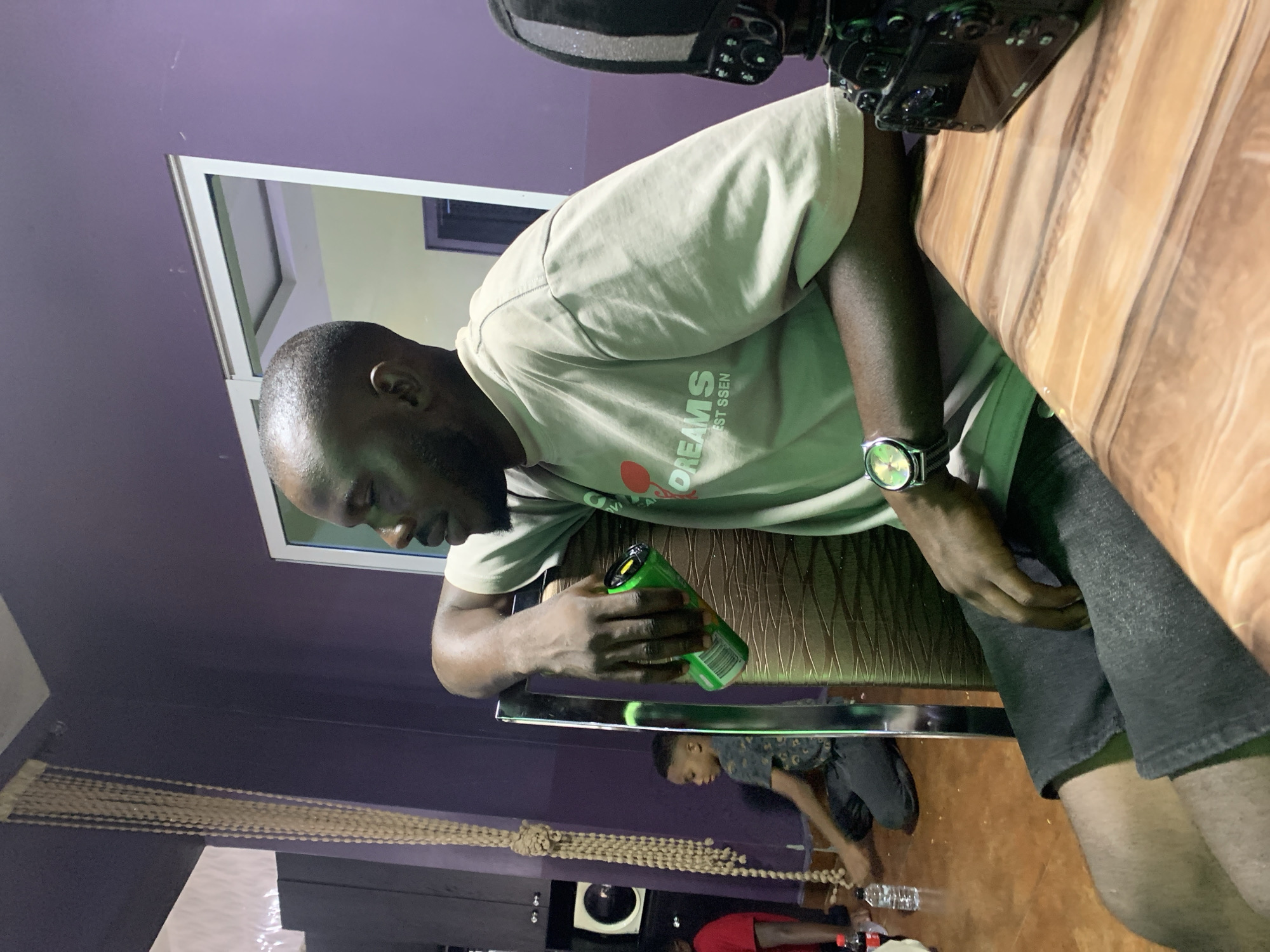 The image depicts an indoor scene in Jos, Nigeria, featuring three individuals in what appears to be a lounge or casual gathering space.
In the foreground and midground, a dark-skinned adult male is seated, leaning against a chair. He has a shaved head and a short beard, wearing a light-colored (possibly beige or light green) short-sleeved t-shirt with the text "DREAMS EST SSEN" and a red graphic on the chest. He also wears dark shorts. His left arm rests on the top of a brown, horizontally textured chair back, holding a green beverage can with a yellow ring pull. A wristwatch with a colorful face and a metallic band is visible on his right wrist. He is looking towards the left side of the frame. To his immediate right in the foreground, on a light brown, wood-grain patterned table, sits a black DSLR camera with an attached flash unit.
In the midground on the left, a dark-skinned child with short hair is kneeling or sitting on the floor, wearing a dark, patterned t-shirt and dark trousers. He is facing towards the right. Partially visible in the background on the far left, behind the child, is another individual wearing a red short-sleeved t-shirt.
The room features matte purple walls. A long, vertical string of light beige, possibly beaded, decoration hangs from the ceiling on the left side. On the floor near the child, a red Coca-Cola bottle and a clear plastic water bottle are present. A white-framed doorway or window is visible on the right wall, offering a view into an adjacent room with light yellow-green walls and a white ceiling featuring recessed lighting. The floor of the primary room is light brown with a visible grain pattern, suggestive of wood or laminate. The lighting appears to be artificial.