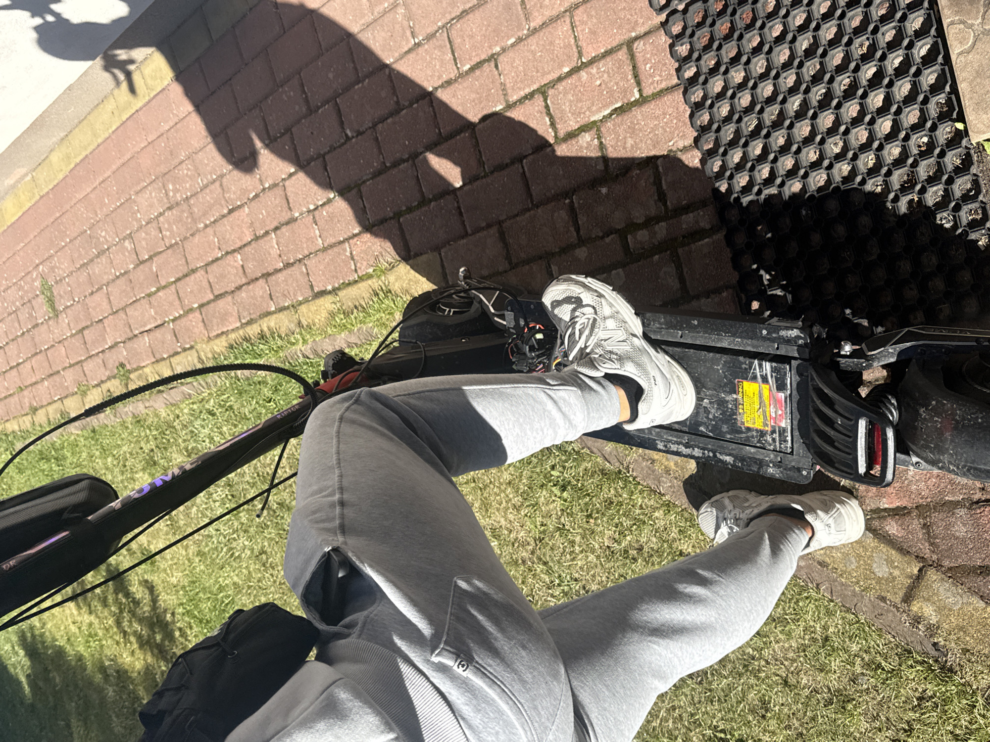 The image shows a person standing on an electric scooter, which is parked on a paved walkway next to a grassy area. The scooter is black and has some wires visible. The person is wearing grey sweatpants and white athletic shoes. A black bag is attached to the handlebars of the scooter. The shadow of a person and the scooter is cast on the walkway, indicating a sunny day. The environment appears to be an outdoor urban or suburban setting, possibly a park or residential area. The mention of Ustronie Morskie, Poland, suggests a possible geographical context for the scene, but there are no specific landmarks visible in the image to confirm this location.