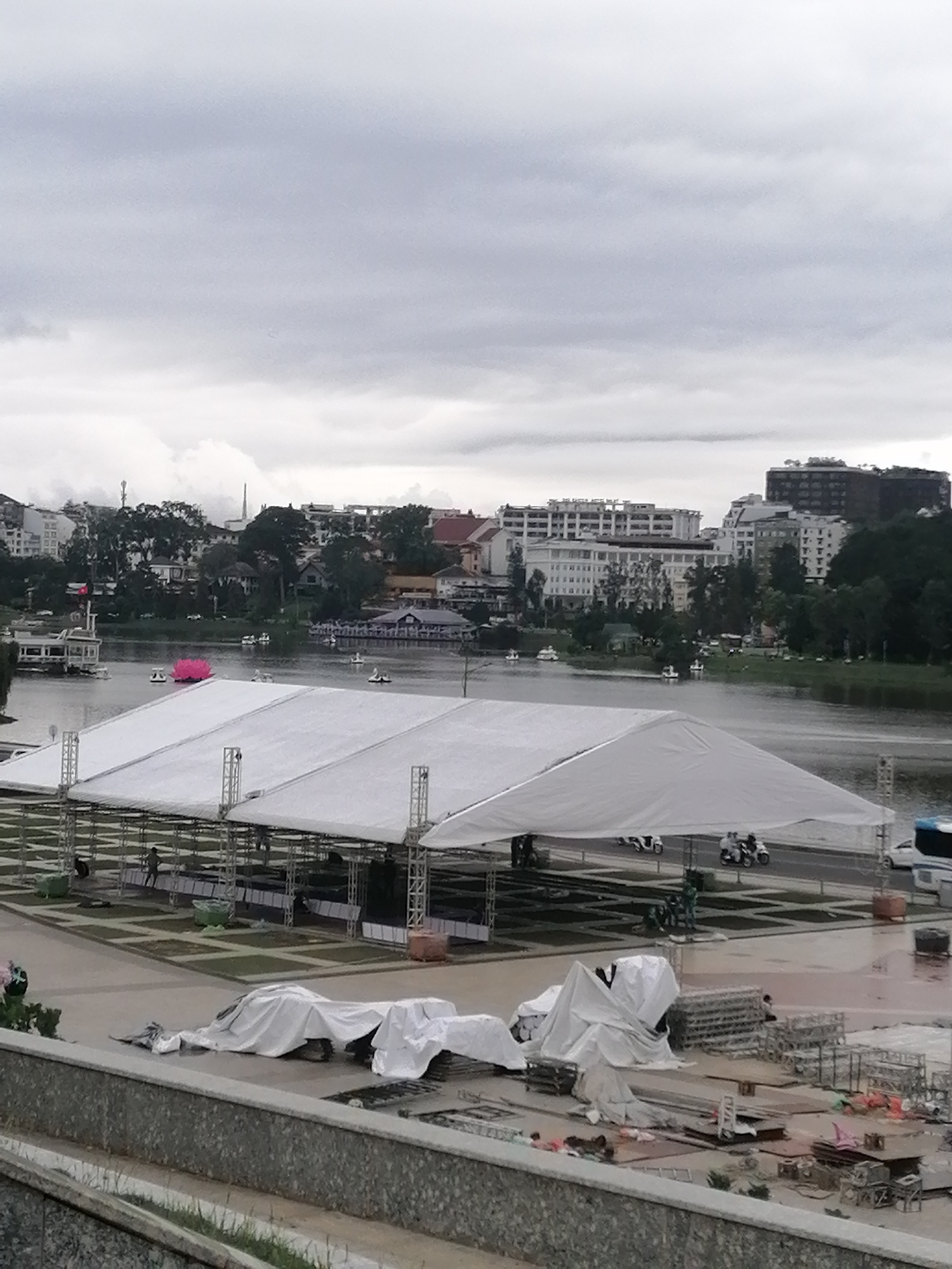 The image captures a cloudy day in Da Lat, Vietnam. In the foreground, construction is underway, with equipment and materials covered in white tarps. A large white tent-like structure dominates the midground, with metal supports visible underneath. People can be seen walking underneath the structure. Behind the tent, a body of water, likely Xuan Huong Lake, reflects the overcast sky. Various watercraft, including swan-shaped paddle boats and a larger boat with a Vietnamese flag, are scattered across the lake. In the background, the cityscape of Da Lat unfolds, with buildings of varying heights and architectural styles interspersed with trees and greenery. The overall impression is one of preparation for an event in a bustling city environment.