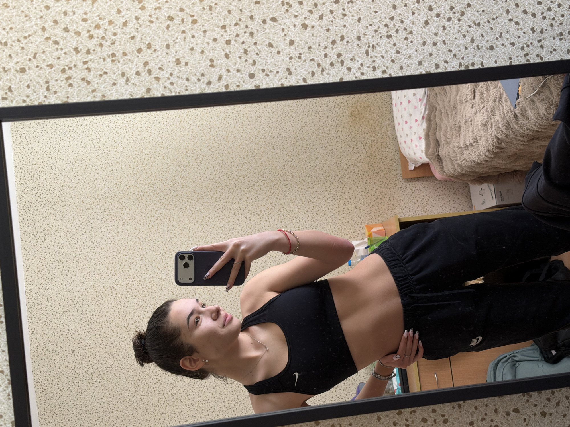 A mirror selfie shows a young woman wearing a black Nike sports bra and black sweatpants. She holds a dark-colored smartphone with a multi-lens camera in her right hand. Her hair is pulled back in a bun. She has bracelets on both wrists and a necklace. The background visible in the mirror shows a room with a speckled wall, a bed with patterned sheets and a textured blanket, and a wooden cabinet. The location cannot be determined based on the image.