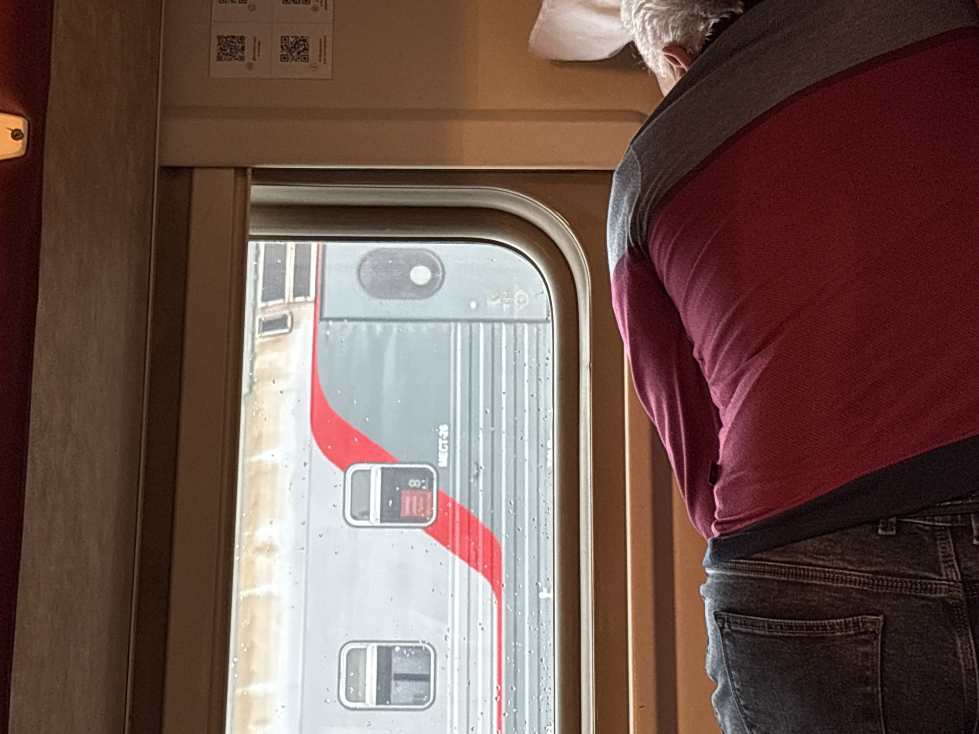 The image shows a person asleep on a train. The person is lying on their side with their back to the camera. They are wearing a red shirt and blue jeans. Outside the window, another train is visible. The scene takes place on a train traveling through Kotelnich, Russia.