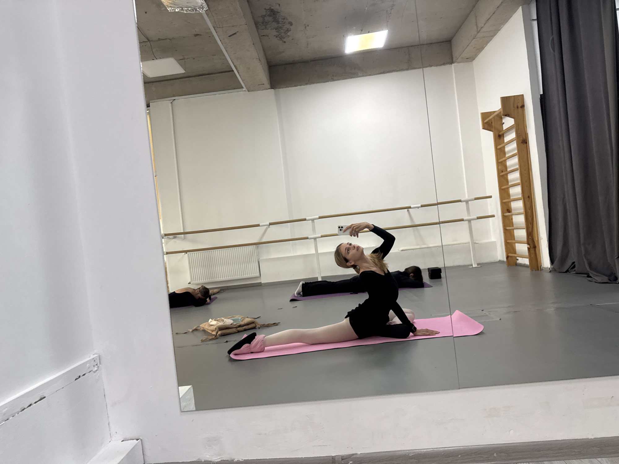 A female ballet dancer in a black leotard and pink tights performs a split on a pink mat in a dance studio. She holds a smartphone up to take a selfie. Two other individuals are visible in the background, one lying down on a mat and another prone on the floor. The studio includes ballet barres, a climbing ladder, and a gray curtain. The location is likely in Tbilisi, Georgia.