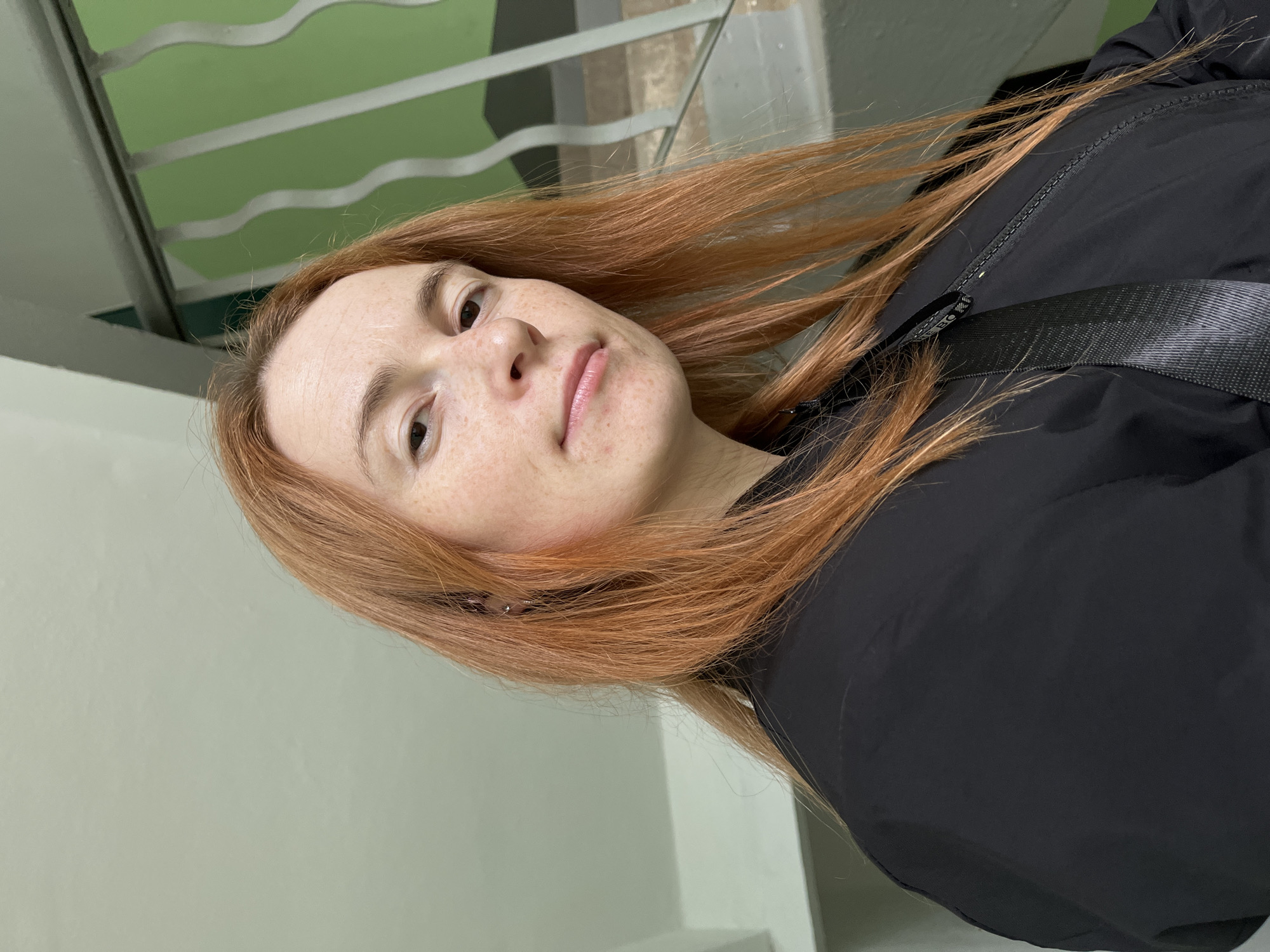 A selfie depicts a fair-skinned woman with red hair, freckles, and a light pink lip color. She is wearing a black jacket with a thick black strap, possibly a backpack or bag strap, across her chest. The background includes a light green wall and what appears to be a white metal railing or decorative element. The location is Chaikovsky, Russia.